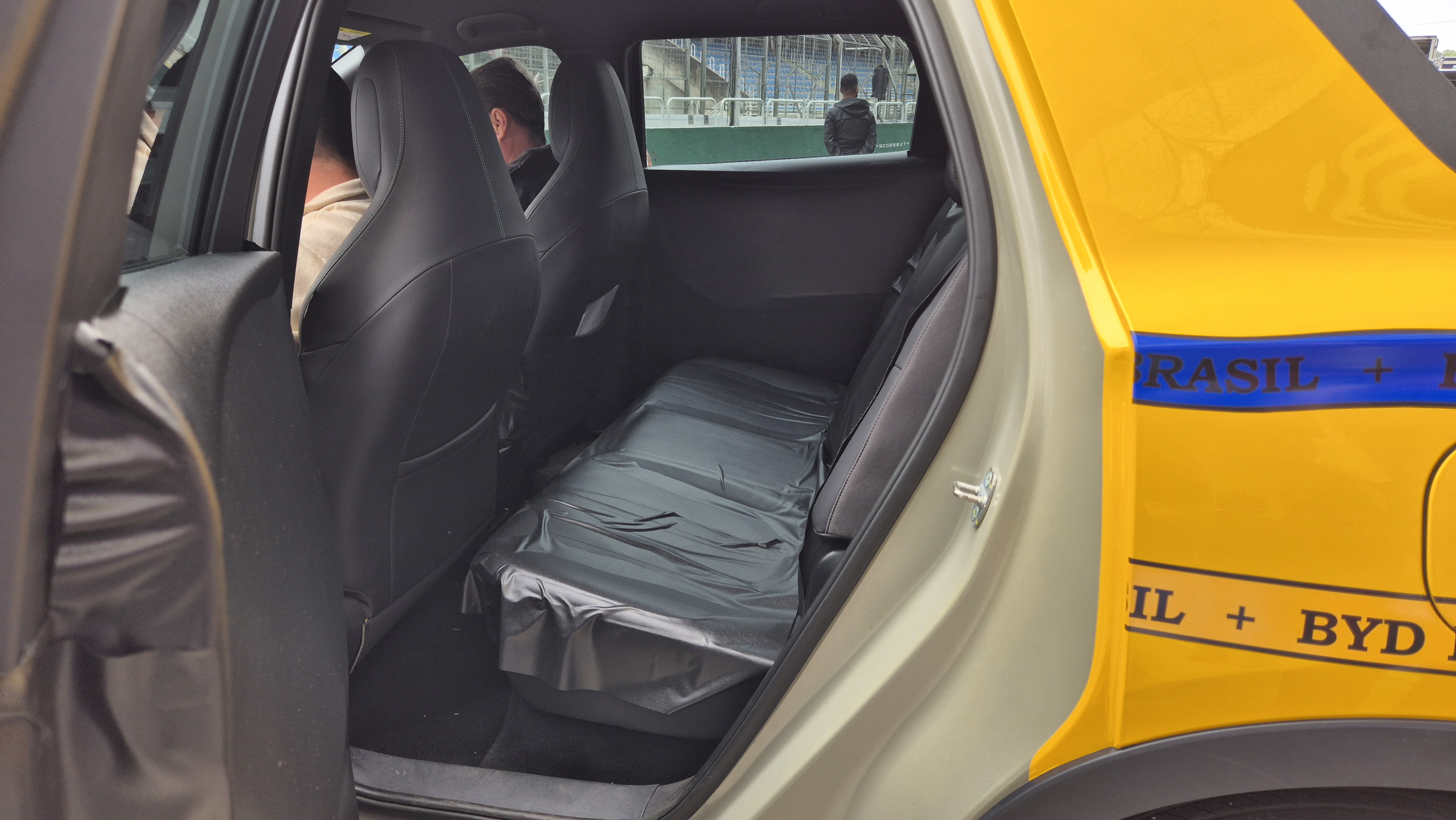 Interior de um carro com bancos pretos, sendo os traseiros cobertos por pl&aacute;stico. Duas pessoas sentadas nos bancos dianteiros, vistas de costas. Pela janela traseira, v&ecirc;-se uma pessoa em p&eacute; e uma &aacute;rea verde. A lateral externa do carro &eacute; amarela com uma faixa azul e o texto BRASIL + BYD em preto