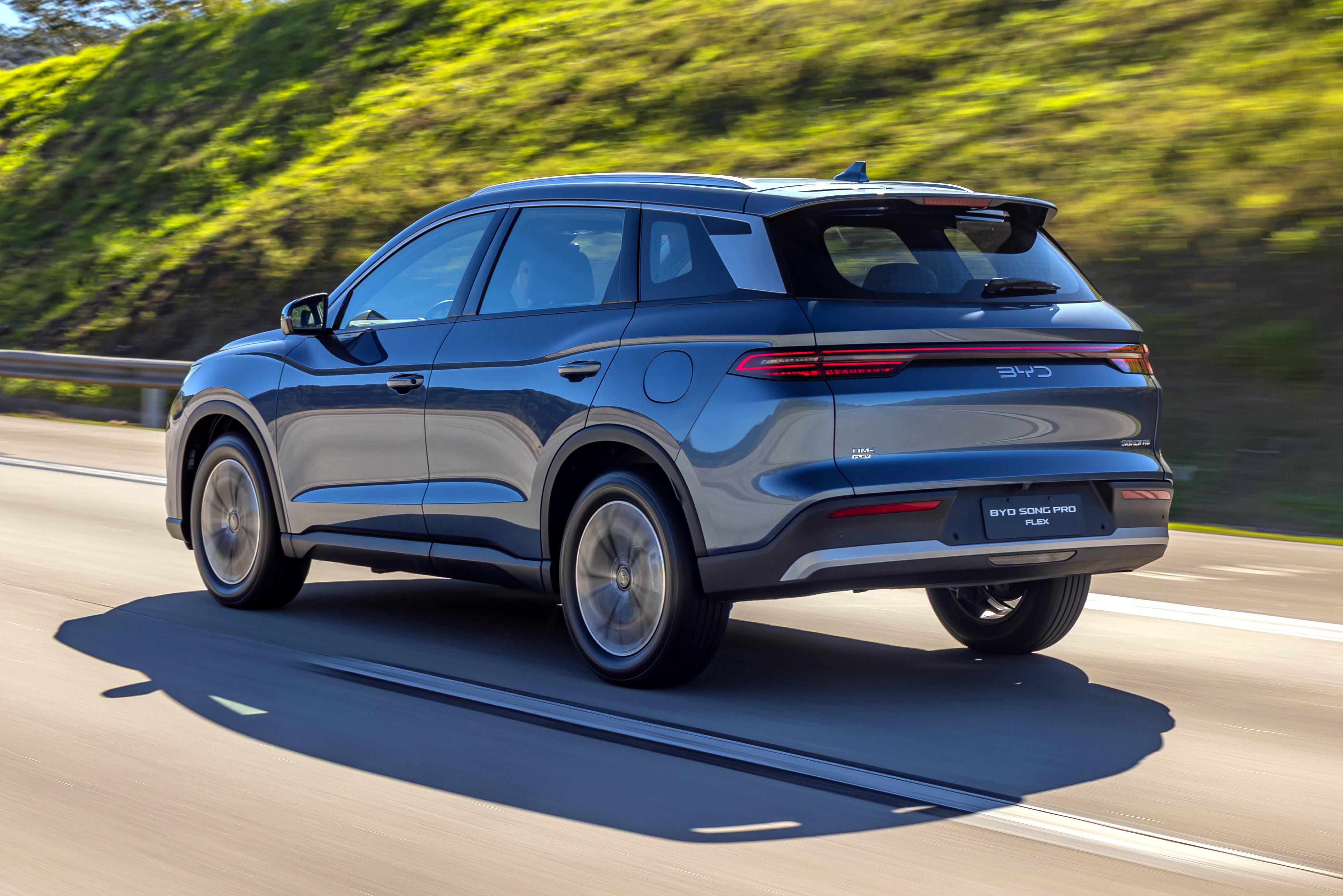 SUV BYD Song Plus DM-i azul escuro em movimento, visto de traseira lateral, em uma estrada com vegeta&ccedil;&atilde;o verde ao fundo
