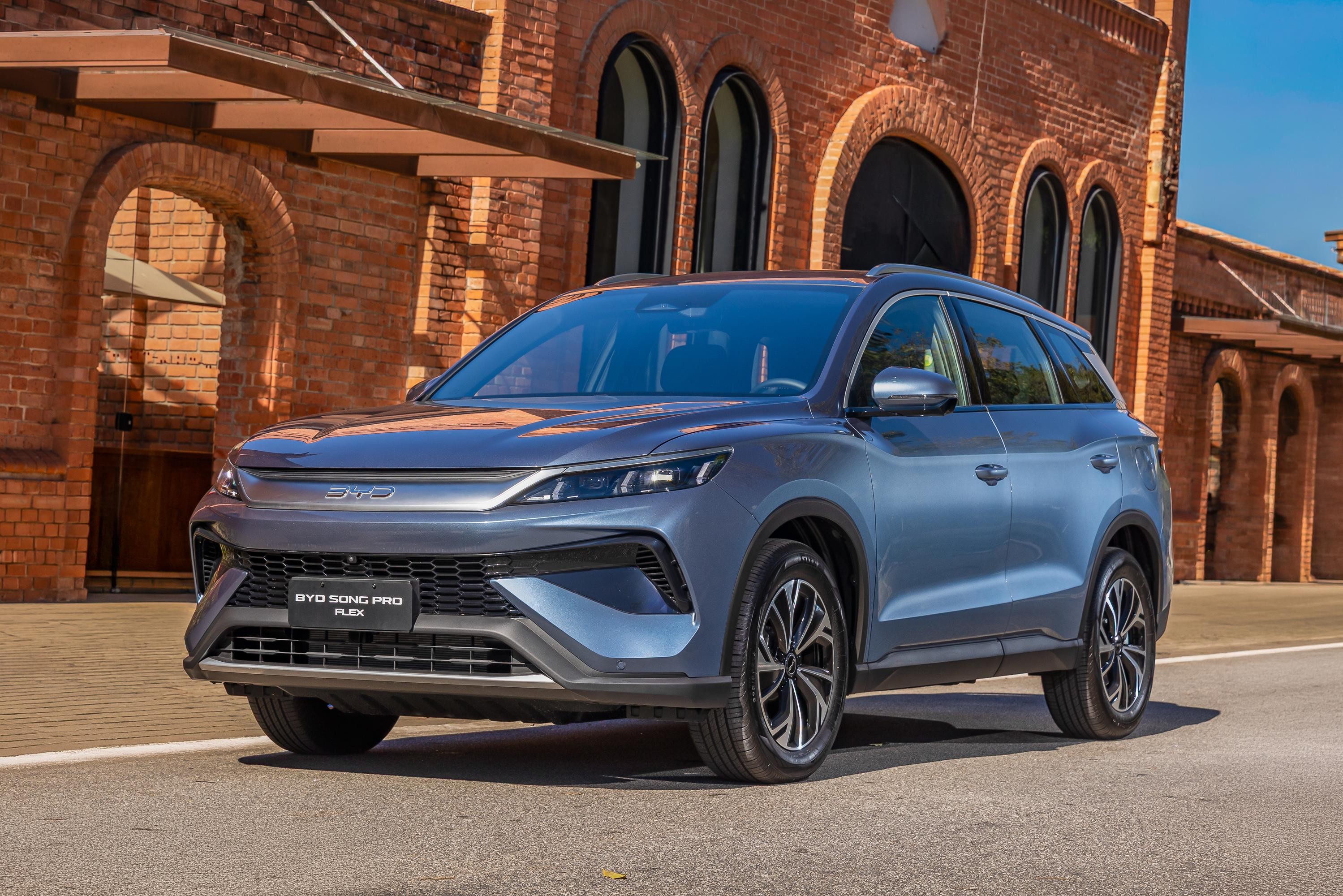 SUV BYD Song Pro Flex azul-claro estacionado em frente a um edif&iacute;cio de tijolos vermelhos com arcos e telhado marrom, sob c&eacute;u azul