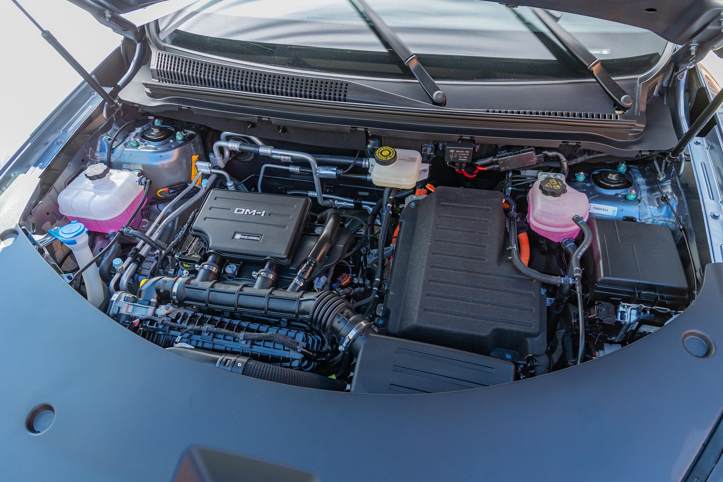 Motor de carro h&iacute;brido com cap&ocirc; aberto, mostrando componentes como reservat&oacute;rios de fluidos rosa e branco, cabos e caixas pretas do sistema el&eacute;trico, e o motor a combust&atilde;o com a inscri&ccedil;&atilde;o DM-i