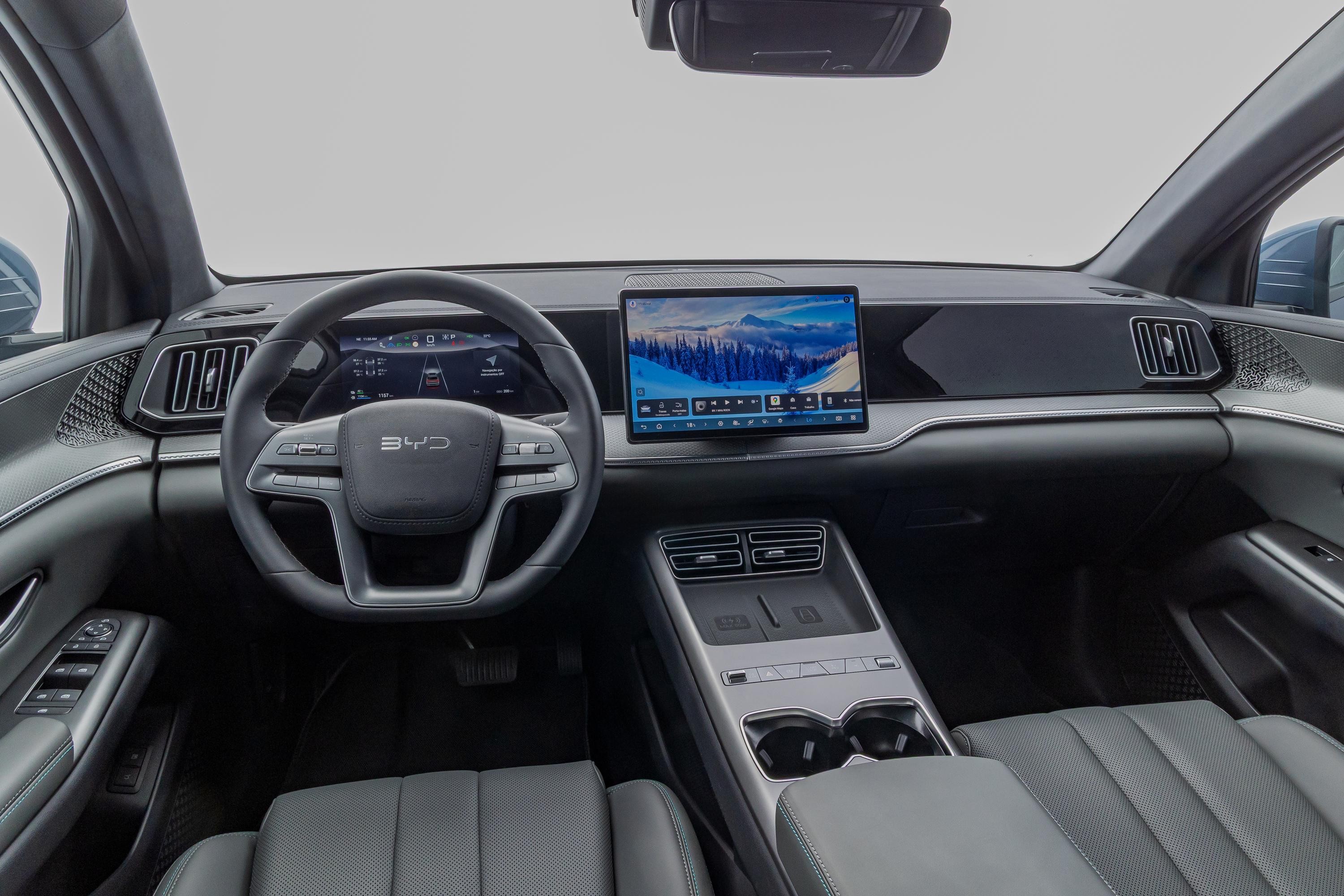 Interior de um carro moderno com volante multifuncional, painel digital e uma tela central grande exibindo uma paisagem de montanhas nevadas. O console central possui porta-copos e carregadores sem fio, e os bancos s&atilde;o de couro cinza escuro