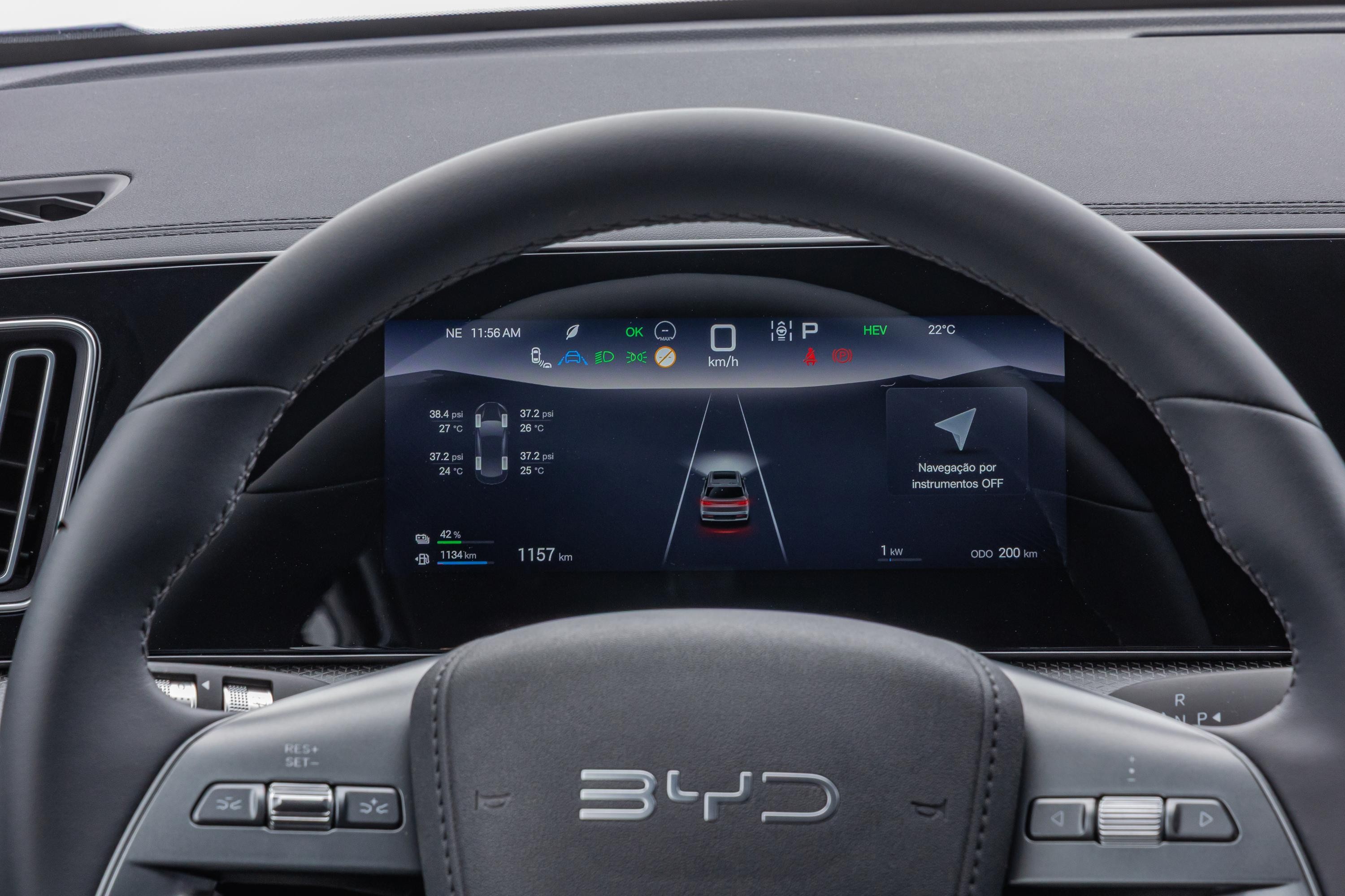 Painel digital de um carro BYD com volante em primeiro plano. O painel exibe informa&ccedil;&otilde;es como hora, temperatura, press&atilde;o dos pneus, n&iacute;vel de bateria, autonomia e um carro vermelho em uma estrada, indicando o sistema de assist&ecirc;ncia ao motorista