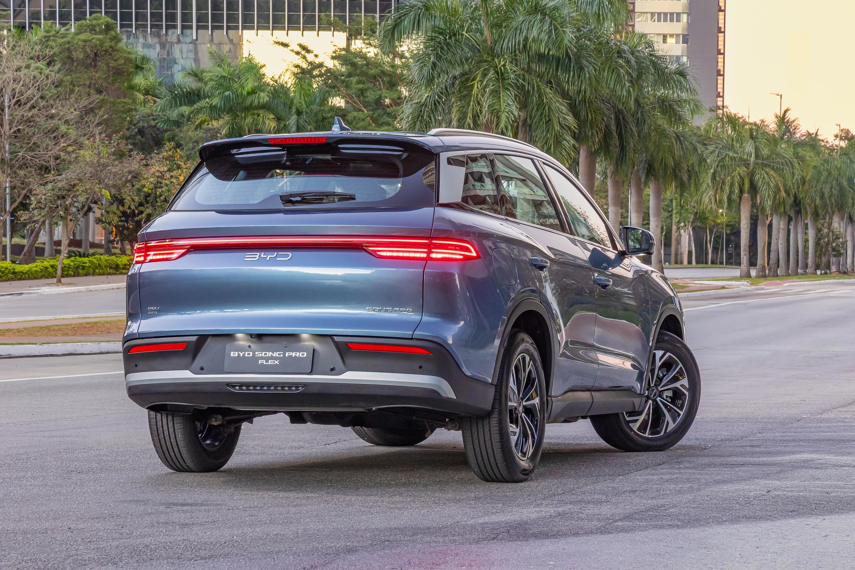 SUV BYD Song Pro Flex azul-escuro, visto de traseira e lateral, parado em rua asfaltada. As lanternas traseiras est&atilde;o acesas, e o para-choque tem detalhes cinzas. Ao fundo, palmeiras e pr&eacute;dios modernos sob c&eacute;u claro