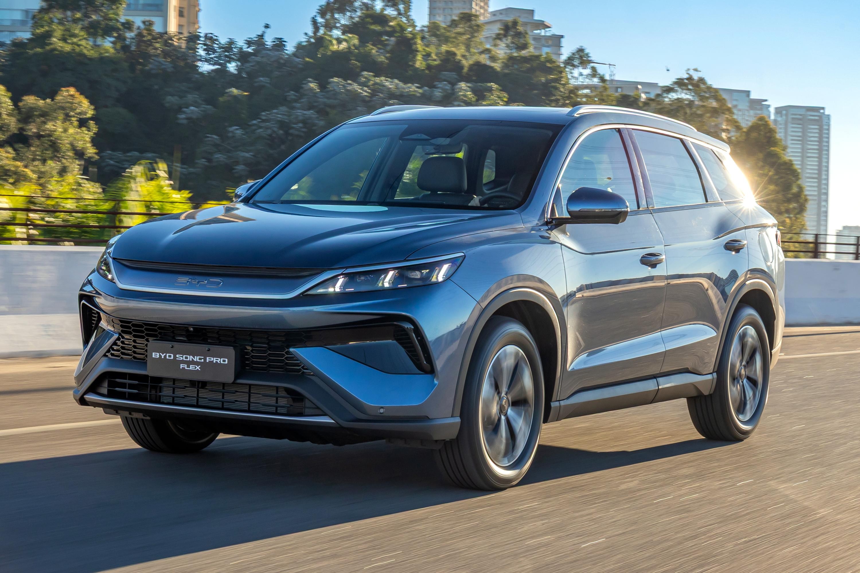 SUV BYD Song Pro Flex azul-escuro em movimento, visto de frente e lateral, com rodas girando. O carro est&aacute; em uma estrada asfaltada, com &aacute;rvores e pr&eacute;dios ao fundo sob um c&eacute;u azul claro. O sol reflete na lateral direita do ve&iacute;culo