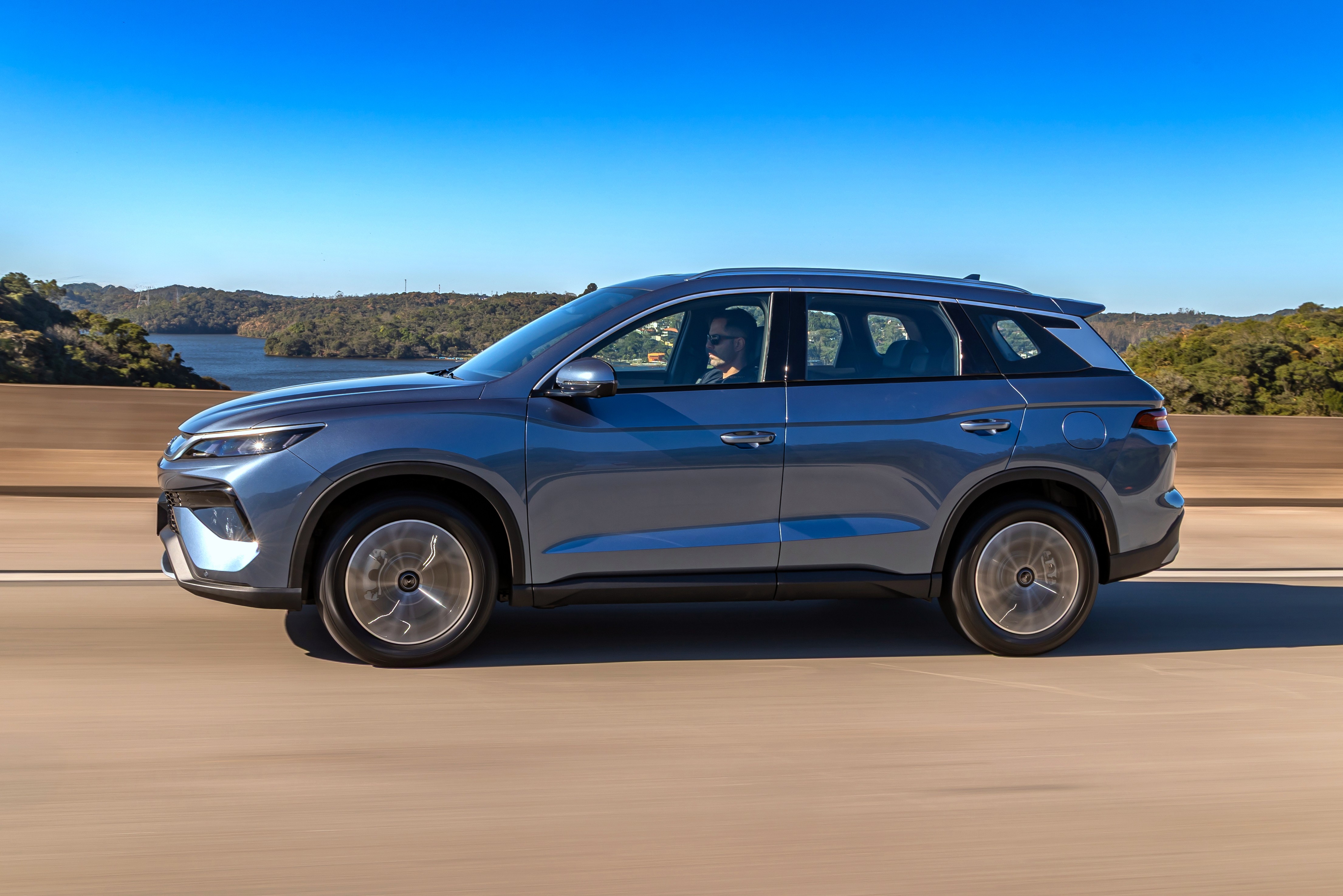SUV azul-acinzentado em movimento na estrada, com um motorista vis&iacute;vel. Ao fundo, um lago cercado por vegeta&ccedil;&atilde;o e um c&eacute;u azul claro