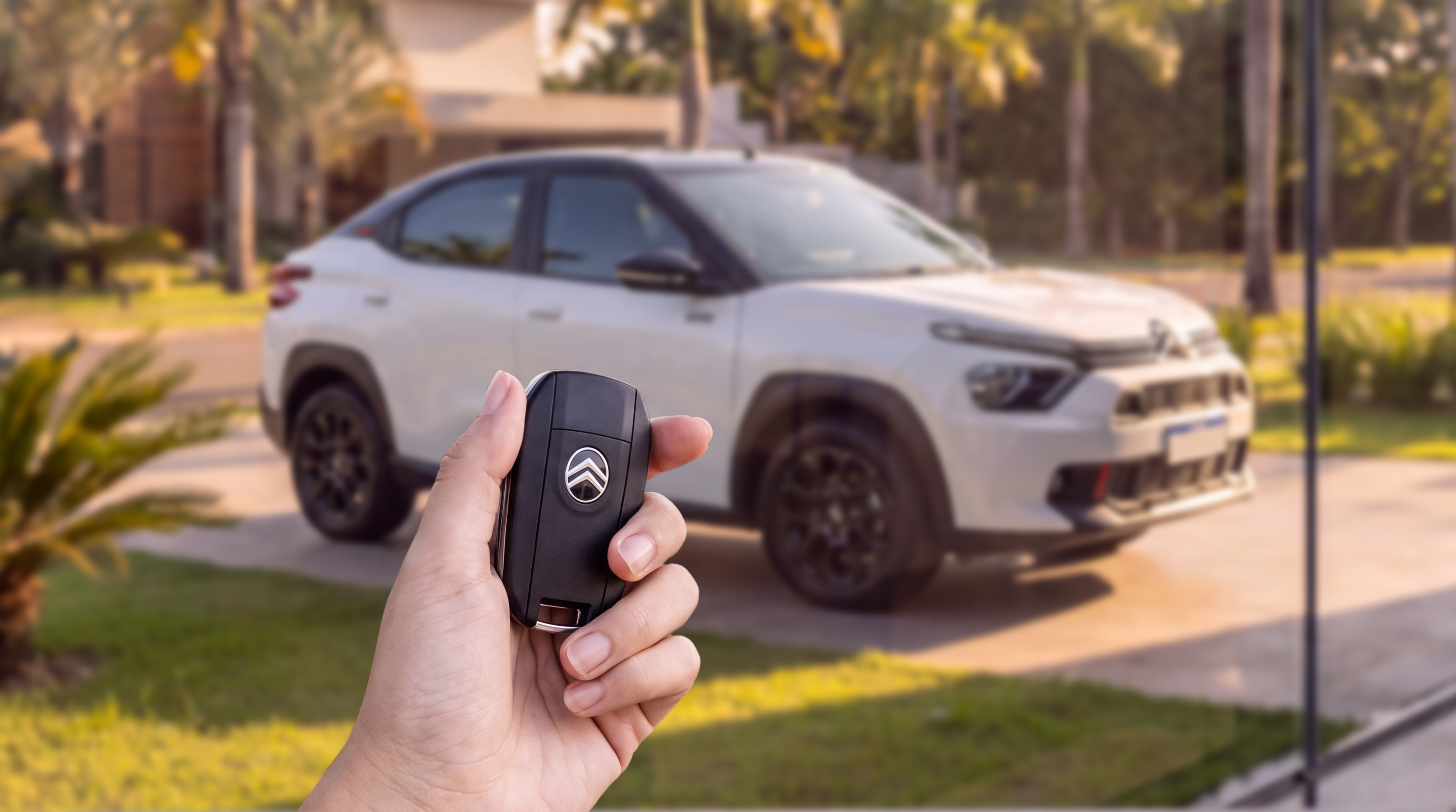 M&atilde;o segurando uma chave de carro preta com o logotipo da Citro&euml;n, com um carro branco e preto desfocado ao fundo, estacionado em uma garagem com gramado e palmeiras