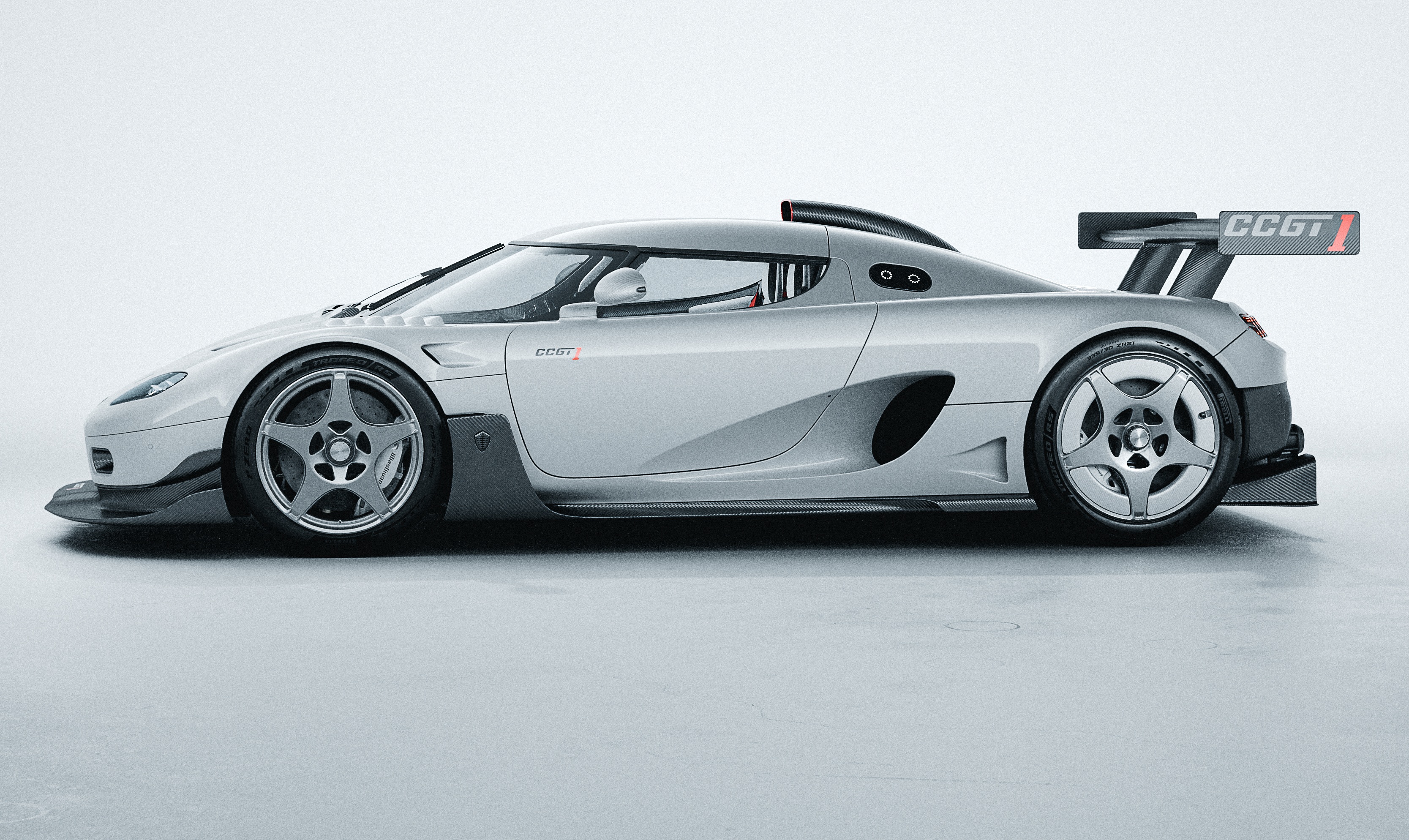 Carro esportivo Koenigsegg CCGT prateado, visto de lado, com aerof&oacute;lio traseiro e rodas de cinco raios, em um est&uacute;dio branco
