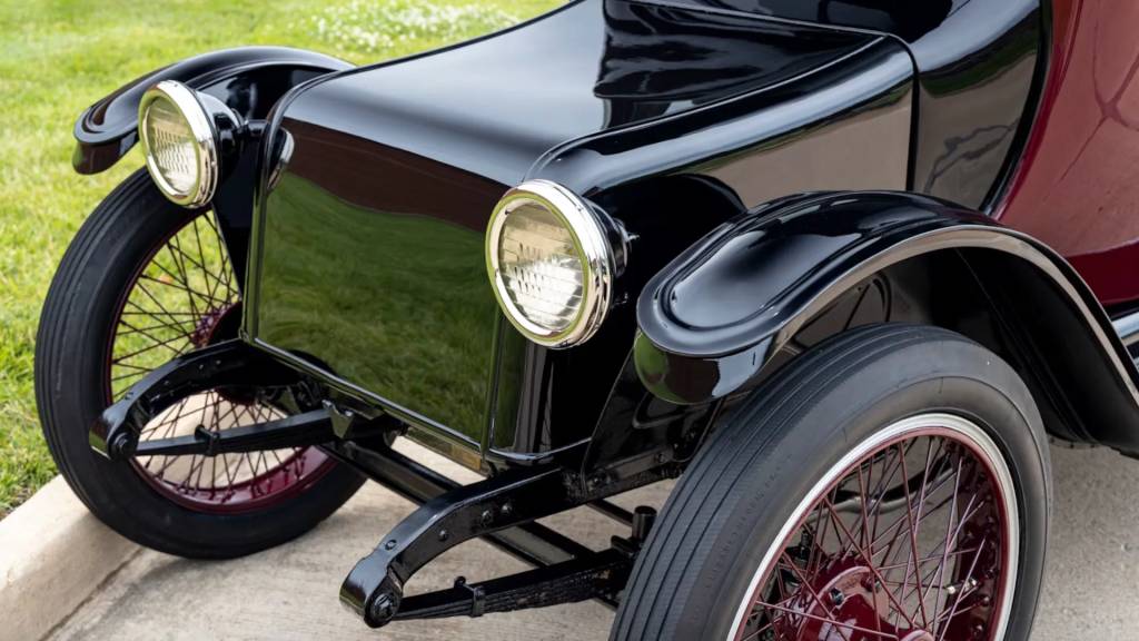 Parte frontal de um carro antigo preto e vinho, com para-lamas e far&oacute;is redondos cromados. As rodas t&ecirc;m aros e raios cor de vinho, e pneus pretos finos. O ve&iacute;culo est&aacute; sobre um piso de concreto, com grama verde ao fundo