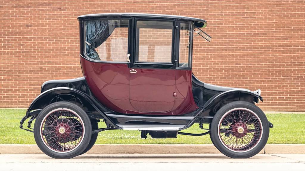 Carro antigo el&eacute;trico de 1914, modelo Detroit Electric, com carroceria preta e portas vinho, rodas raiadas com detalhes vermelhos, estacionado em frente a uma parede de tijolos e gramado