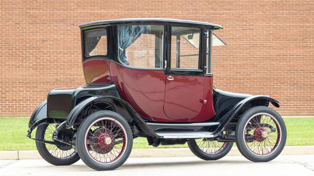 Carro antigo el&eacute;trico, modelo Detroit Electric, com carroceria vinho e para-lamas pretos, rodas de raios finos com aros e cubos vermelhos, estacionado em frente a uma parede de tijolos marrons e gramado verde
