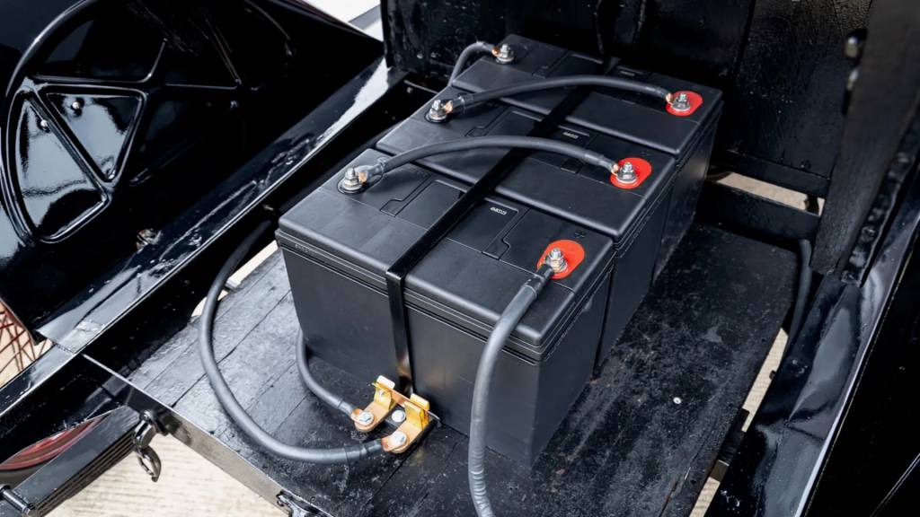 Tr&ecirc;s baterias automotivas pretas retangulares conectadas por cabos grossos, com terminais vermelhos e prateados, instaladas em um compartimento escuro de um ve&iacute;culo antigo