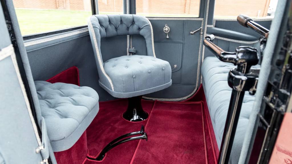 Interior de um carro antigo com estofamento cinza-azulado e carpete vermelho. H&aacute; tr&ecirc;s assentos vis&iacute;veis: um girat&oacute;rio no centro, um menor &agrave; esquerda e um banco comprido &agrave; direita. As paredes s&atilde;o cinza-escuras com detalhes cromados e uma janela retangular. O volante e painel est&atilde;o parcialmente vis&iacute;veis &agrave; direita