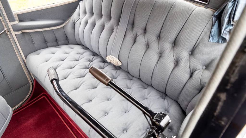Interior de um carro antigo com banco traseiro estofado em tecido cinza claro, com bot&otilde;es e costuras formando losangos. O assoalho &eacute; coberto por um tapete vermelho e duas alavancas pretas com detalhes cromados e marrons se estendem do lado direito. Uma faixa de tecido com franjas brancas pende do encosto do banco.