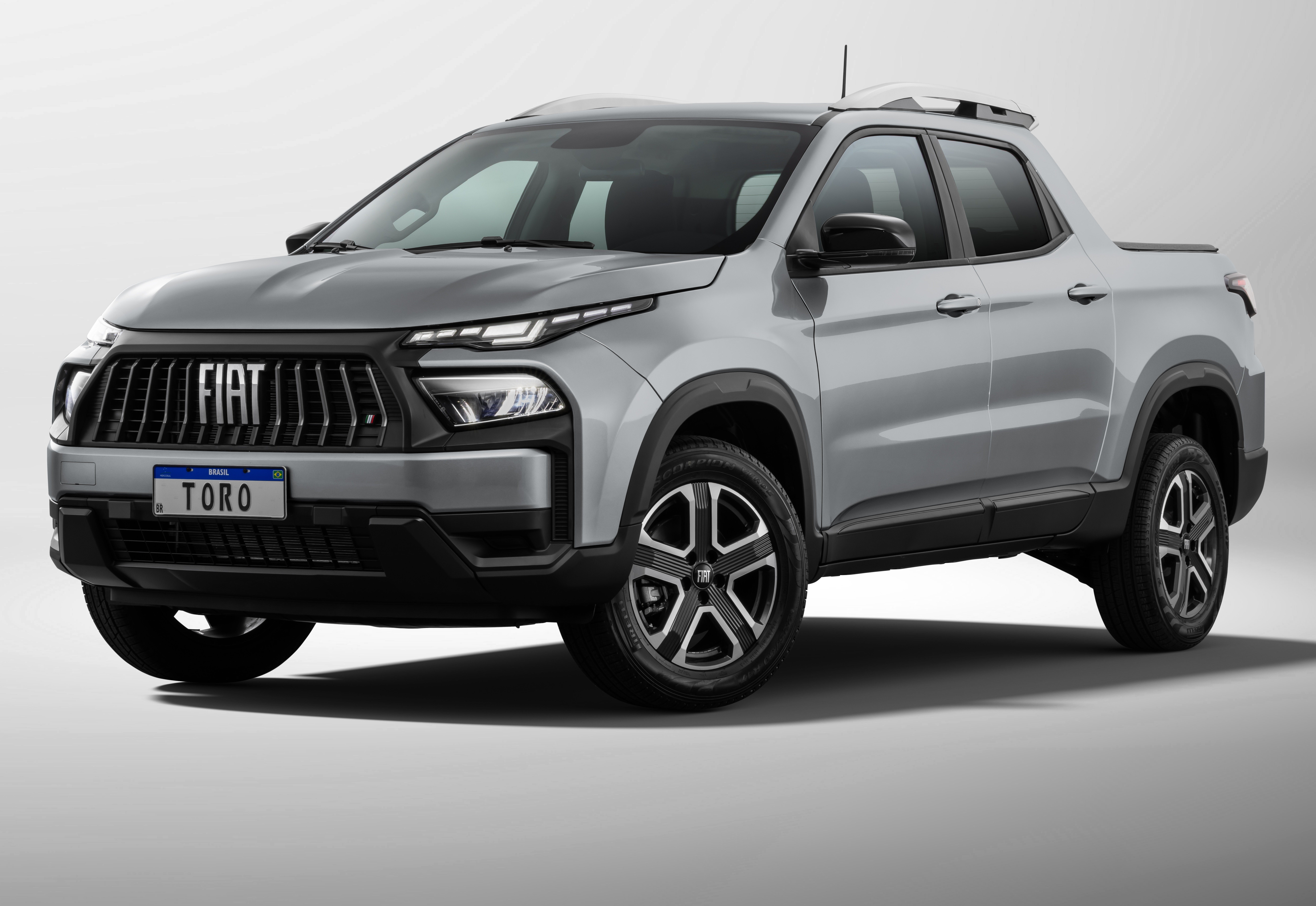 Picape Fiat Toro cinza-claro, com grade frontal preta e far&oacute;is de LED afilados, estacionada em um fundo branco. A placa frontal exibe TORO