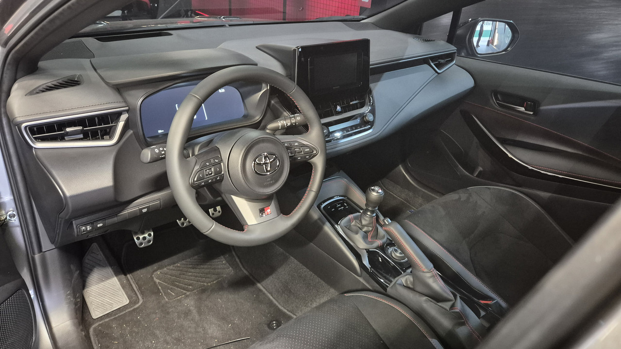 Interior de um carro esportivo cinza escuro, com volante multifuncional e logo Toyota, detalhes vermelhos no volante e c&acirc;mbio manual. O painel possui tela digital e central multim&iacute;dia, bancos de tecido escuro e pedais de metal. A porta do motorista est&aacute; aberta, revelando o retrovisor lateral.