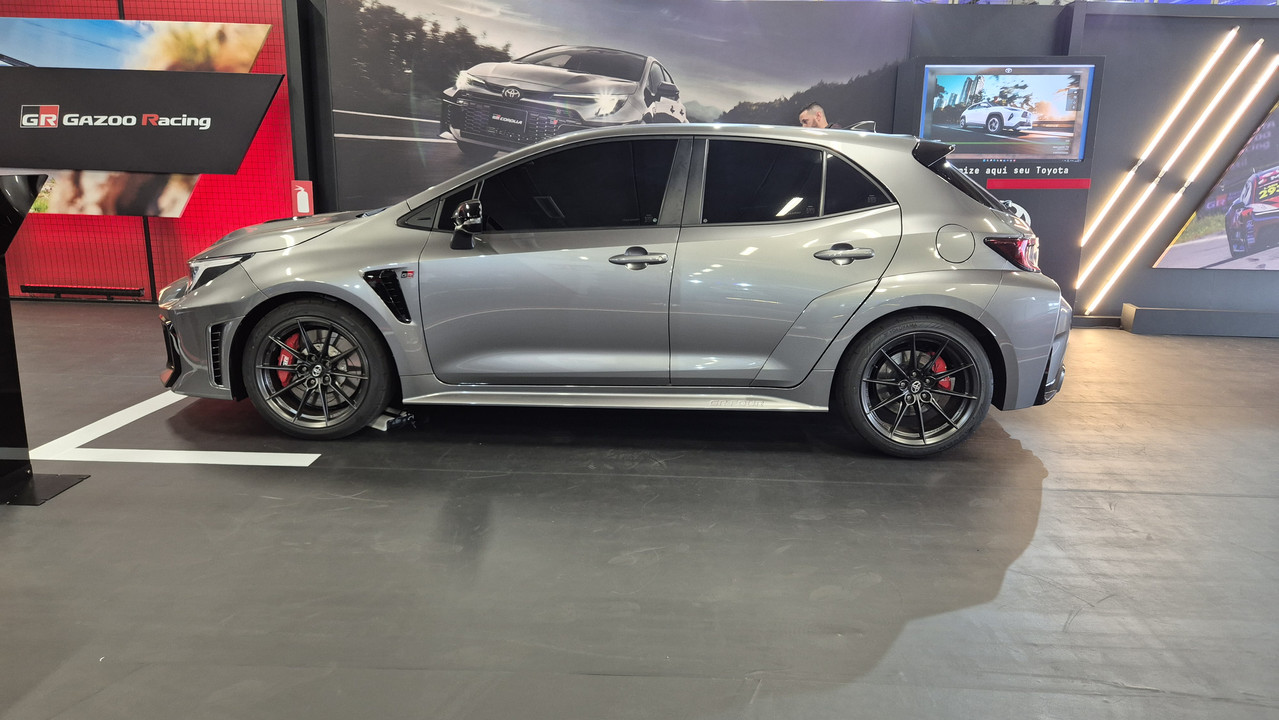 Carro esportivo cinza-chumbo, modelo hatchback, com rodas pretas e pin&ccedil;as de freio vermelhas, estacionado em um showroom com pain&eacute;is de fundo exibindo outros ve&iacute;culos e o logo GR Gazoo Racing