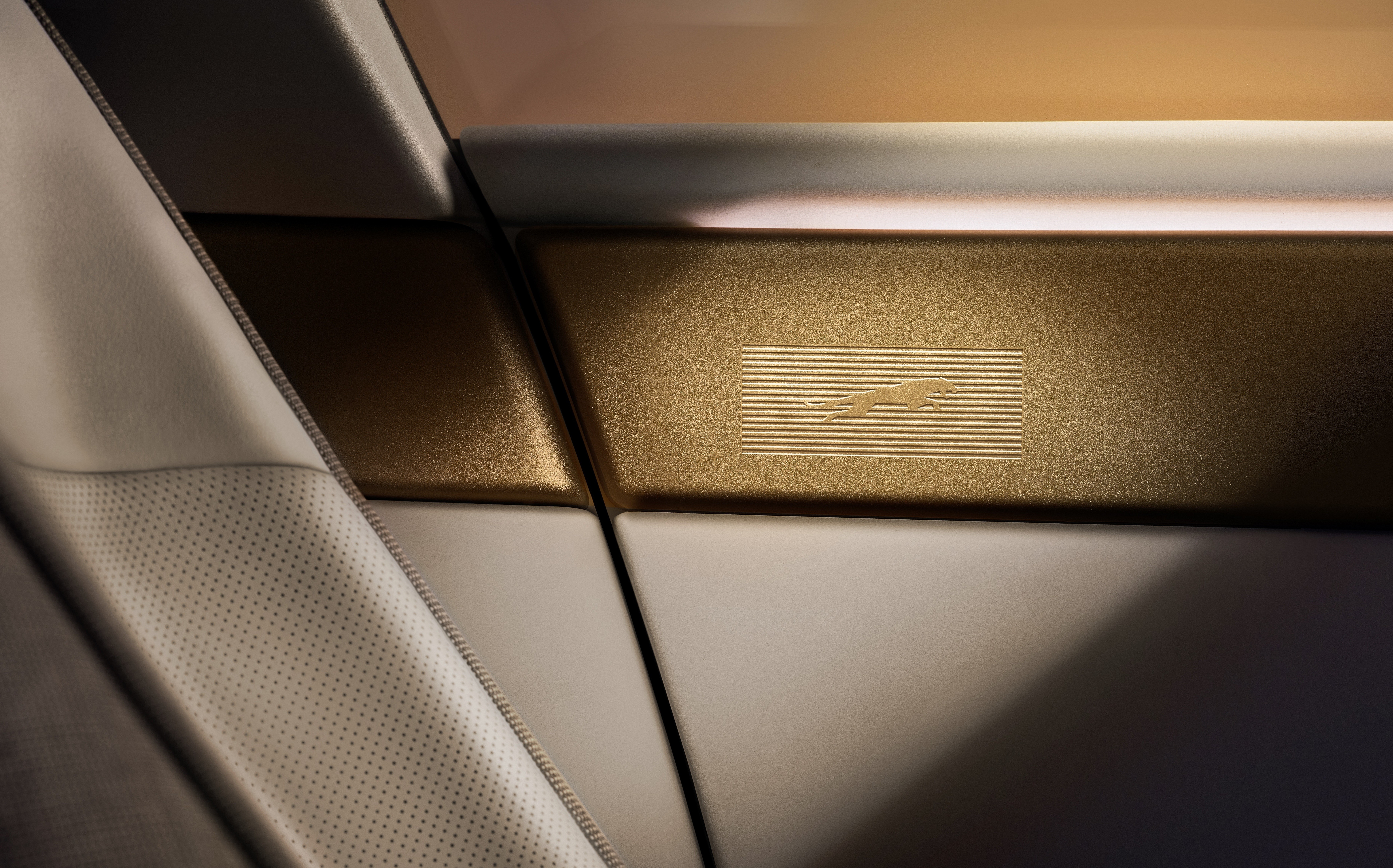 Detalhe do interior de um carro com acabamento em tons de branco e dourado. &Agrave; esquerda, um assento de couro branco perfurado. No centro, uma faixa horizontal dourada com o logotipo de um jaguar em relevo sobre linhas horizontais finas. A ilumina&ccedil;&atilde;o cria um contraste de luz e sombra, destacando a textura dos materiais