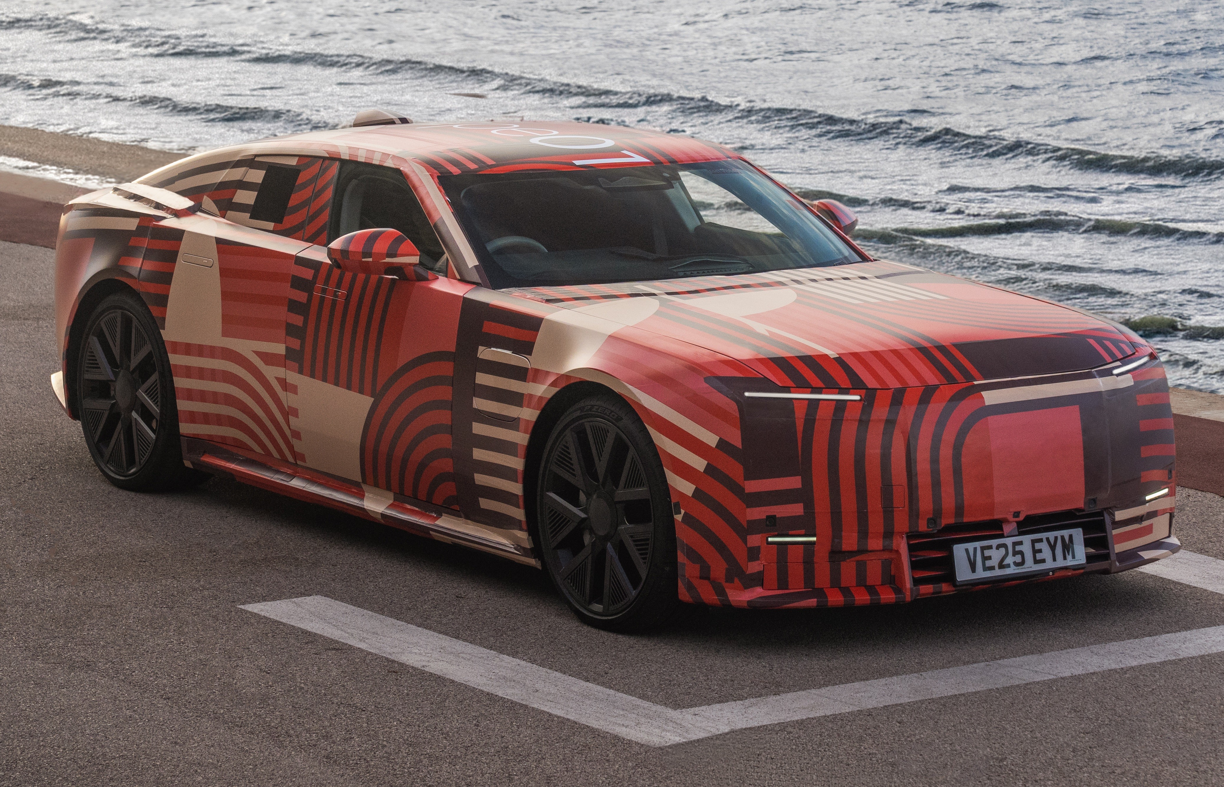 Carro prot&oacute;tipo camuflado com padr&otilde;es geom&eacute;tricos em tons de vermelho, bege e preto, estacionado em uma rua asfaltada com o mar ao fundo
