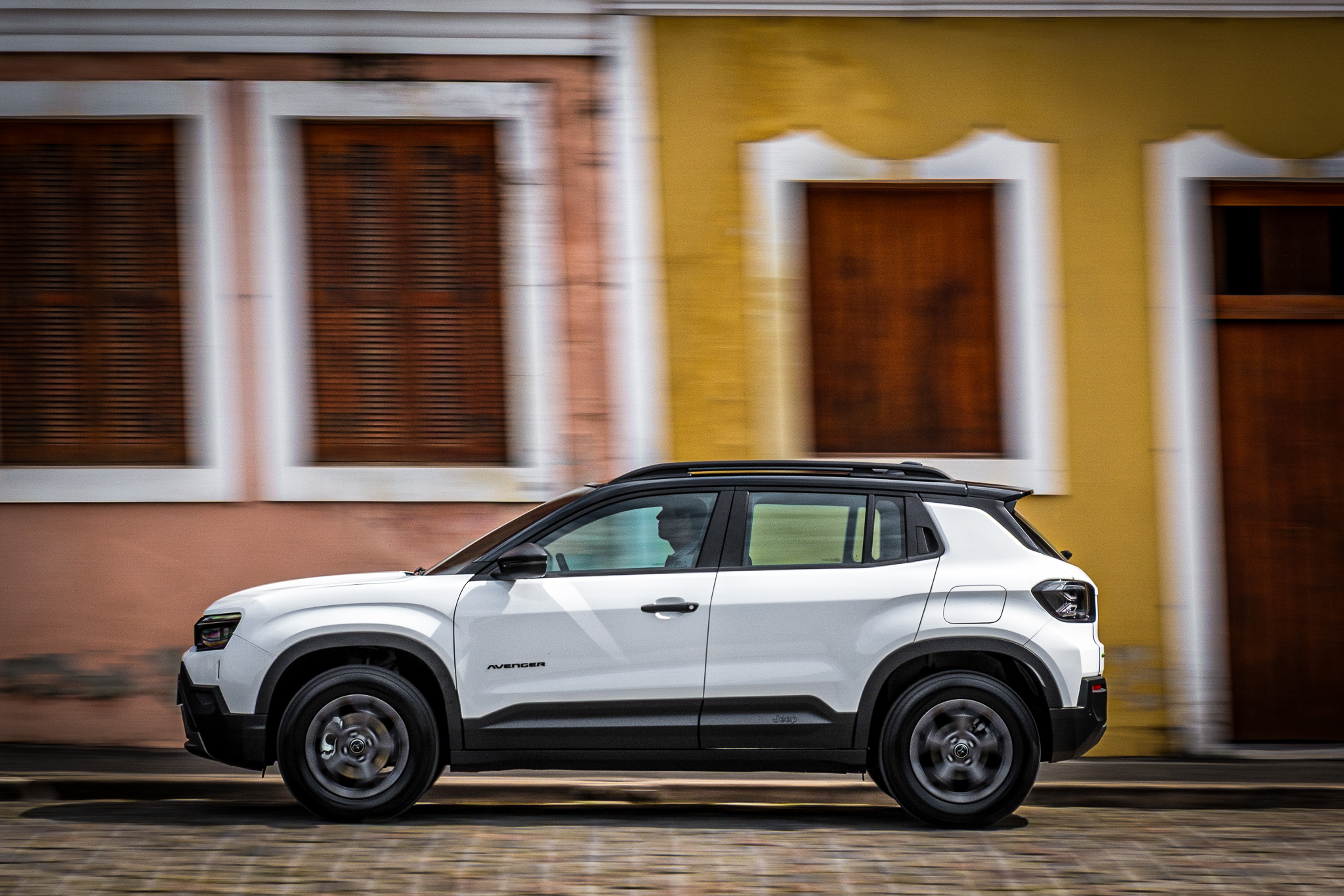 SUV branco com teto preto em movimento, visto de lado, passando por uma rua de paralelep&iacute;pedos. Ao fundo, edif&iacute;cios coloridos com janelas e portas de madeira escura, borrados pelo movimento