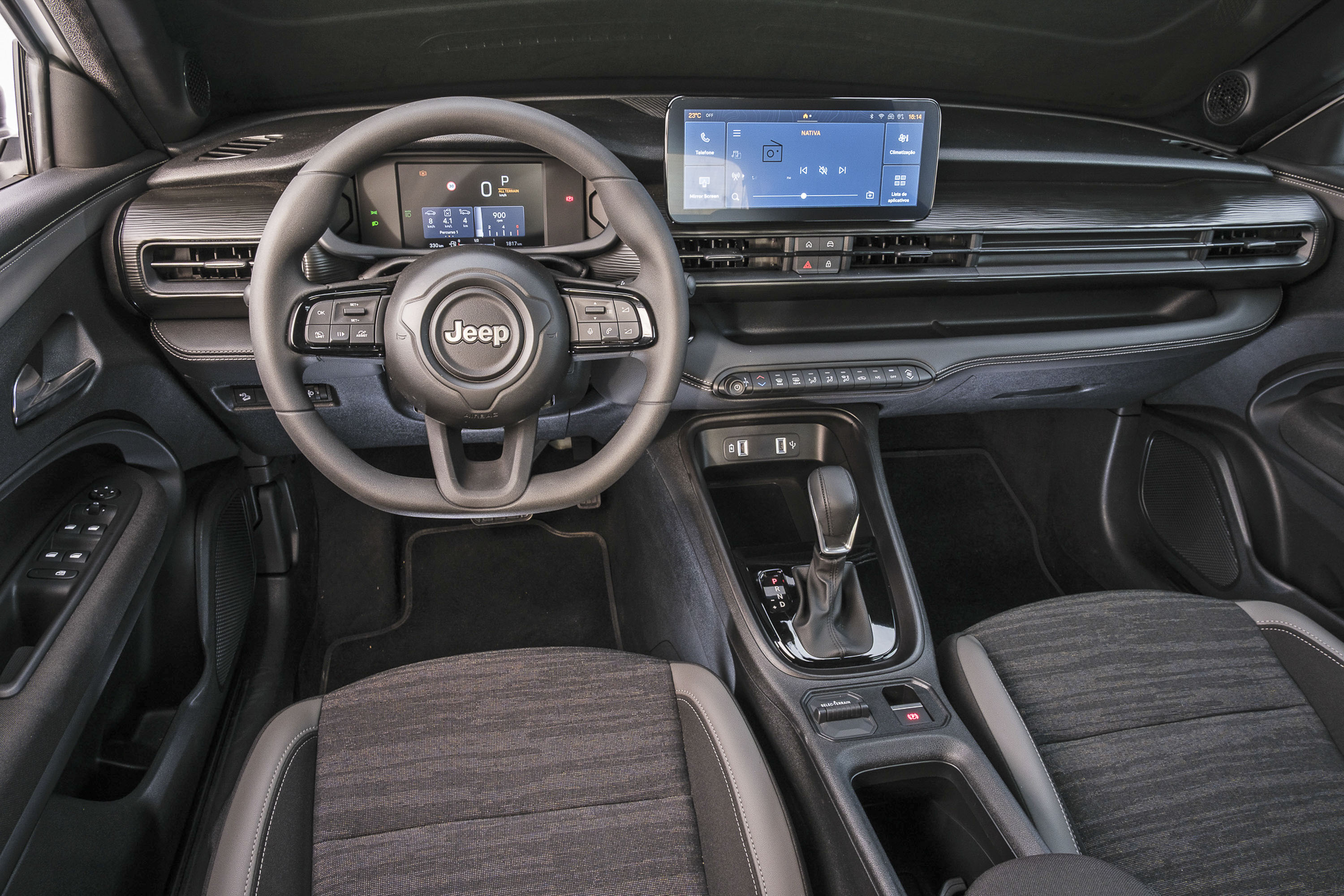 Interior de um carro Jeep, mostrando o volante com o logo da marca, painel digital, tela multim&iacute;dia central com interface azul, console central com c&acirc;mbio autom&aacute;tico e bancos de tecido escuro com detalhes cinza
