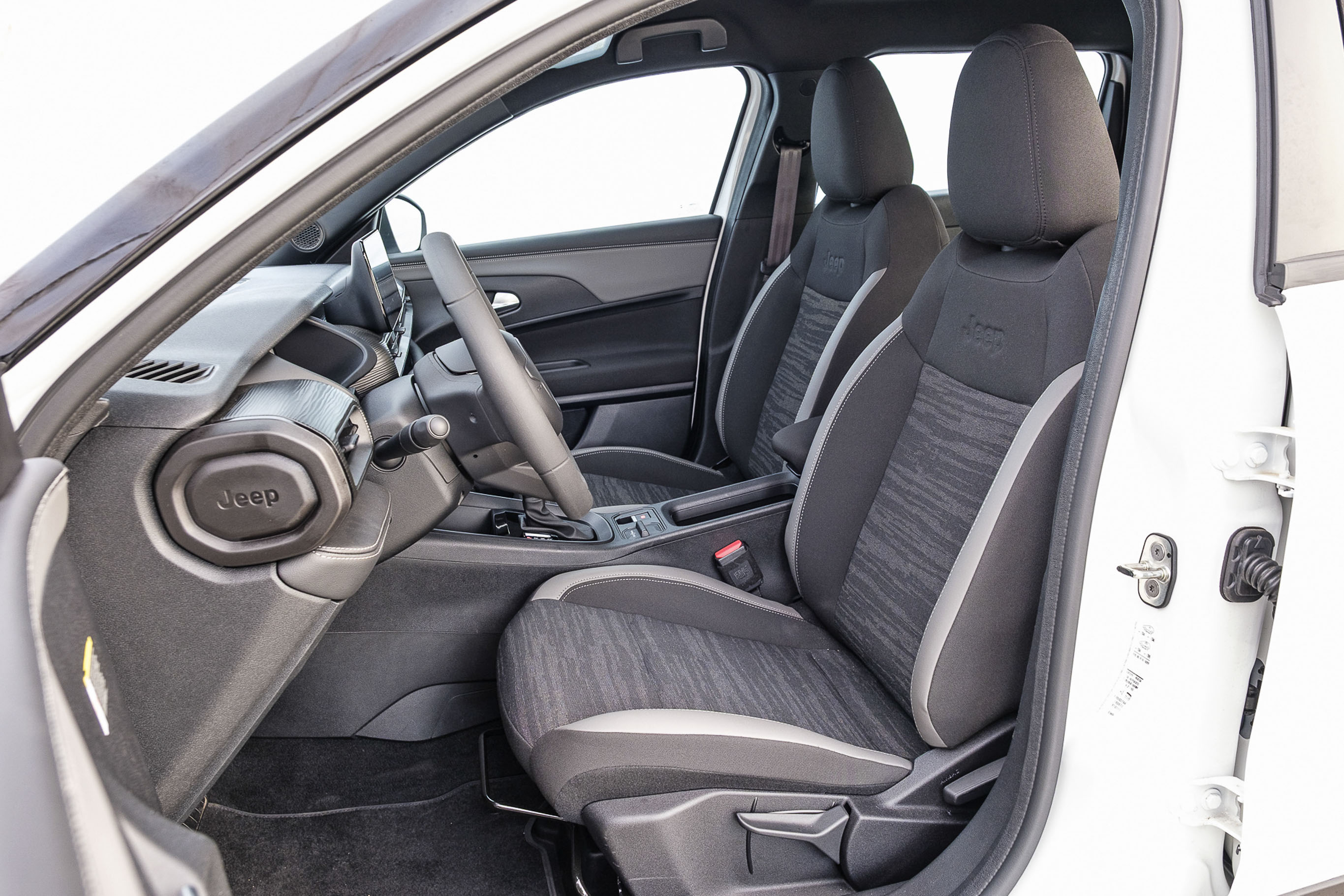 Interior de um carro branco, mostrando o banco do motorista e do passageiro da frente em tecido cinza escuro com detalhes em cinza claro e estampa abstrata. O volante e o painel s&atilde;o pretos, com o logotipo Jeep vis&iacute;vel no painel lateral esquerdo. A porta do motorista est&aacute; aberta, revelando o batente branco