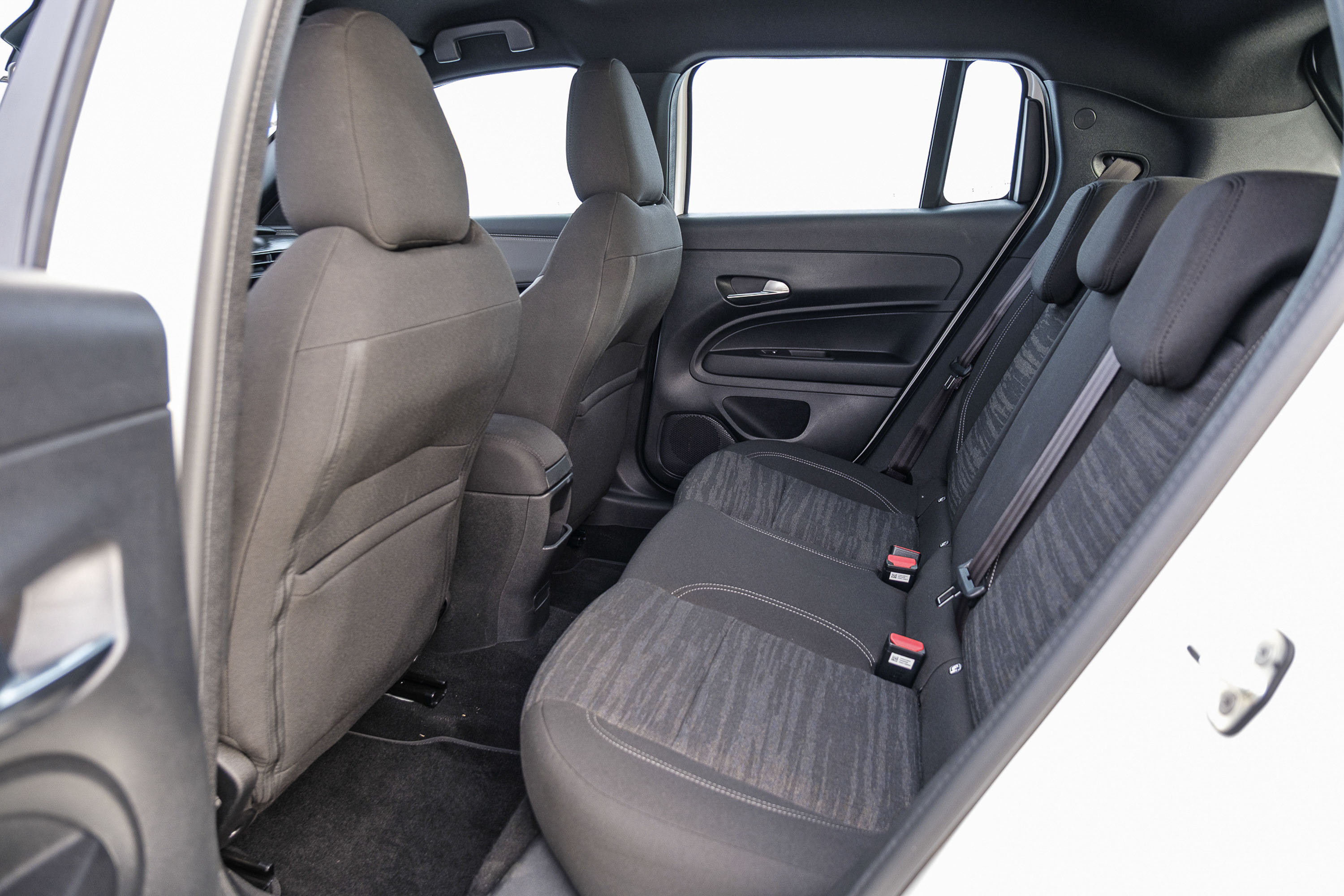 Interior de um carro branco, mostrando os bancos traseiros e parte dos dianteiros. Os bancos s&atilde;o cinza escuro, com estofamento de tecido texturizado nos assentos traseiros e encostos de cabe&ccedil;a pretos. Cintos de seguran&ccedil;a com fivelas vermelhas est&atilde;o vis&iacute;veis. As portas e o teto s&atilde;o pretos, e as janelas mostram um fundo branco claro. A imagem &eacute; tirada de dentro do carro, com a porta traseira direita aberta.