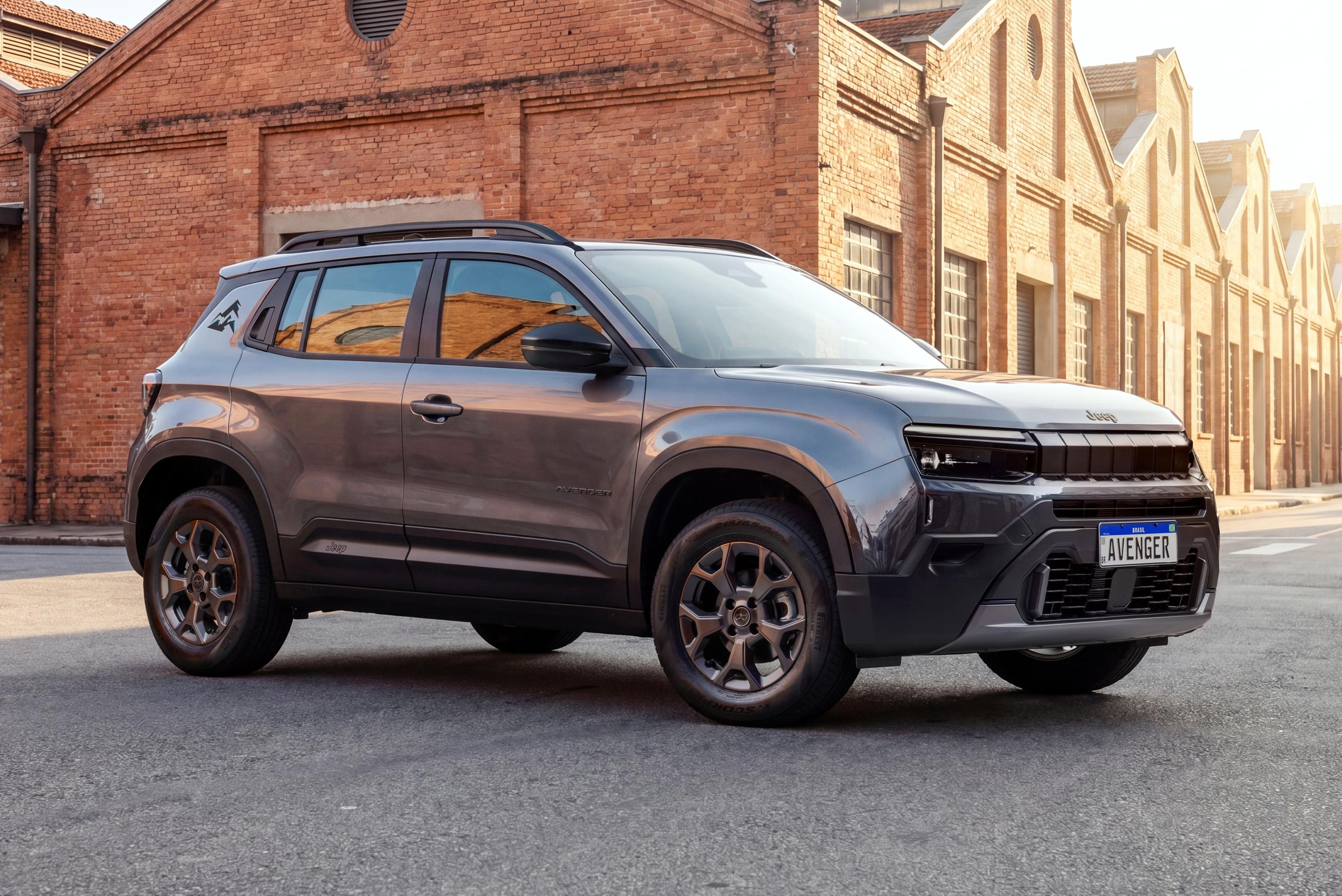 SUV Jeep Avenger cinza escuro estacionado em rua de paralelep&iacute;pedos, com pr&eacute;dios de tijolos avermelhados ao fundo e luz solar forte &agrave; direita