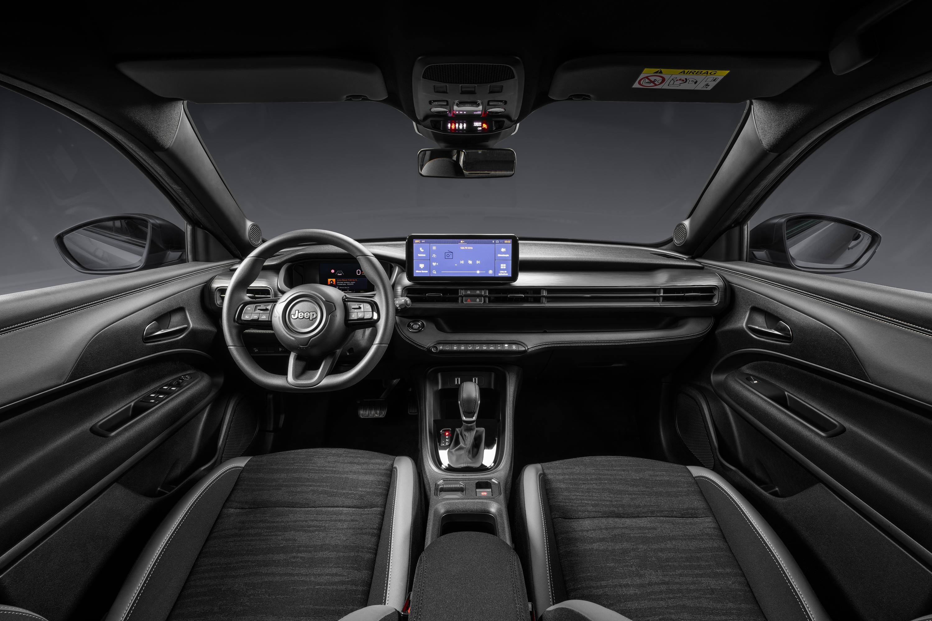 Interior de um carro Jeep moderno, com painel e estofamento em tons escuros. Volante multifuncional com o logo Jeep, tela multim&iacute;dia central, sa&iacute;das de ar horizontais e console com alavanca de c&acirc;mbio. Os bancos dianteiros s&atilde;o de tecido cinza escuro com detalhes em couro.