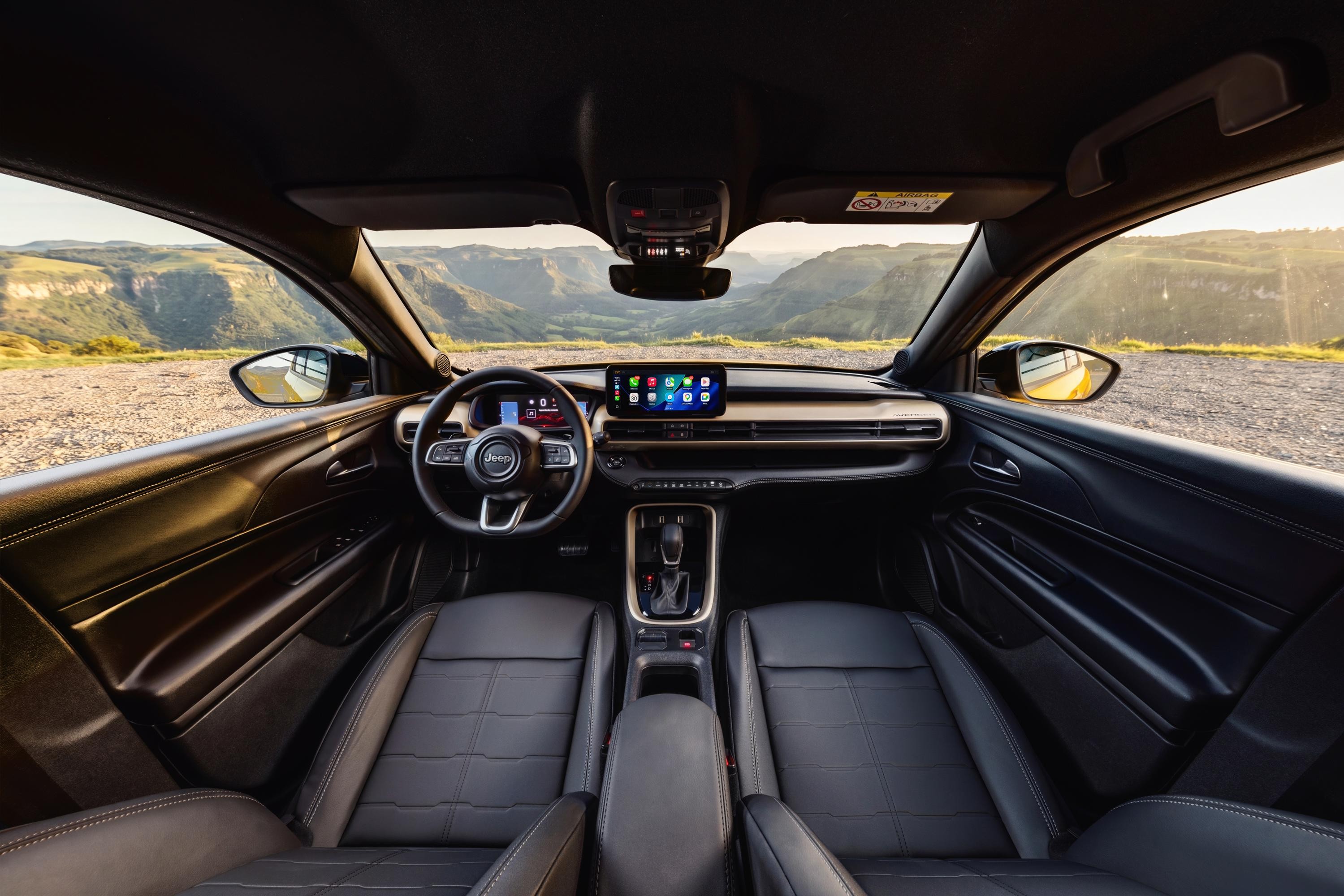 Interior de um carro Jeep preto, com bancos de couro, volante multifuncional e tela multim&iacute;dia central exibindo &iacute;cones de aplicativos. O painel tem detalhes em tom dourado. A vista atrav&eacute;s do para-brisa e das janelas laterais mostra uma paisagem montanhosa com c&eacute;u claro e sol poente, iluminando a estrada de terra. Os retrovisores externos refletem a paisagem.