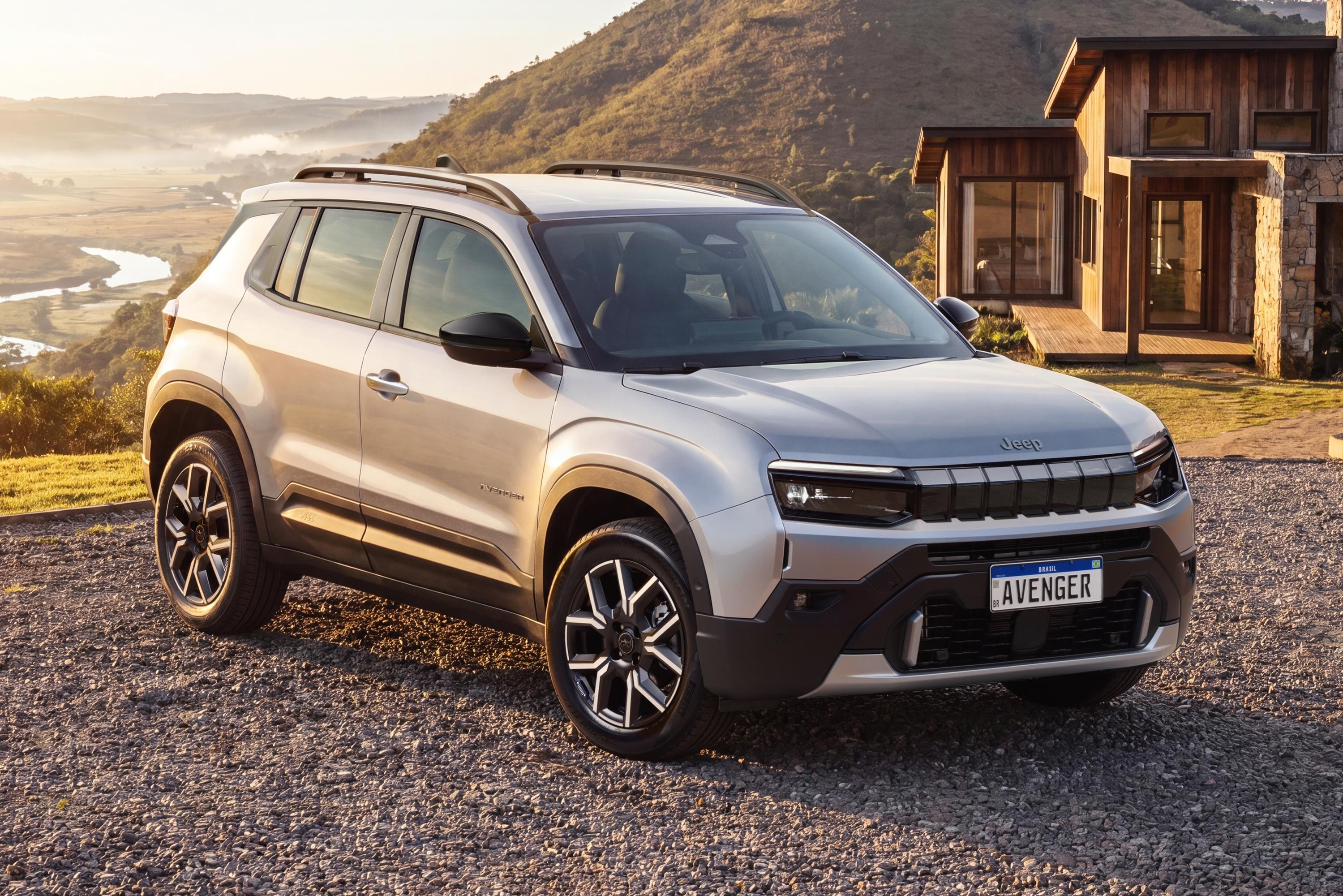 SUV Jeep Avenger prateado estacionado em estrada de cascalho, com casa de madeira e pedra ao fundo. &Agrave; esquerda, um vale com rio sinuoso e montanhas sob o sol da manh&atilde;