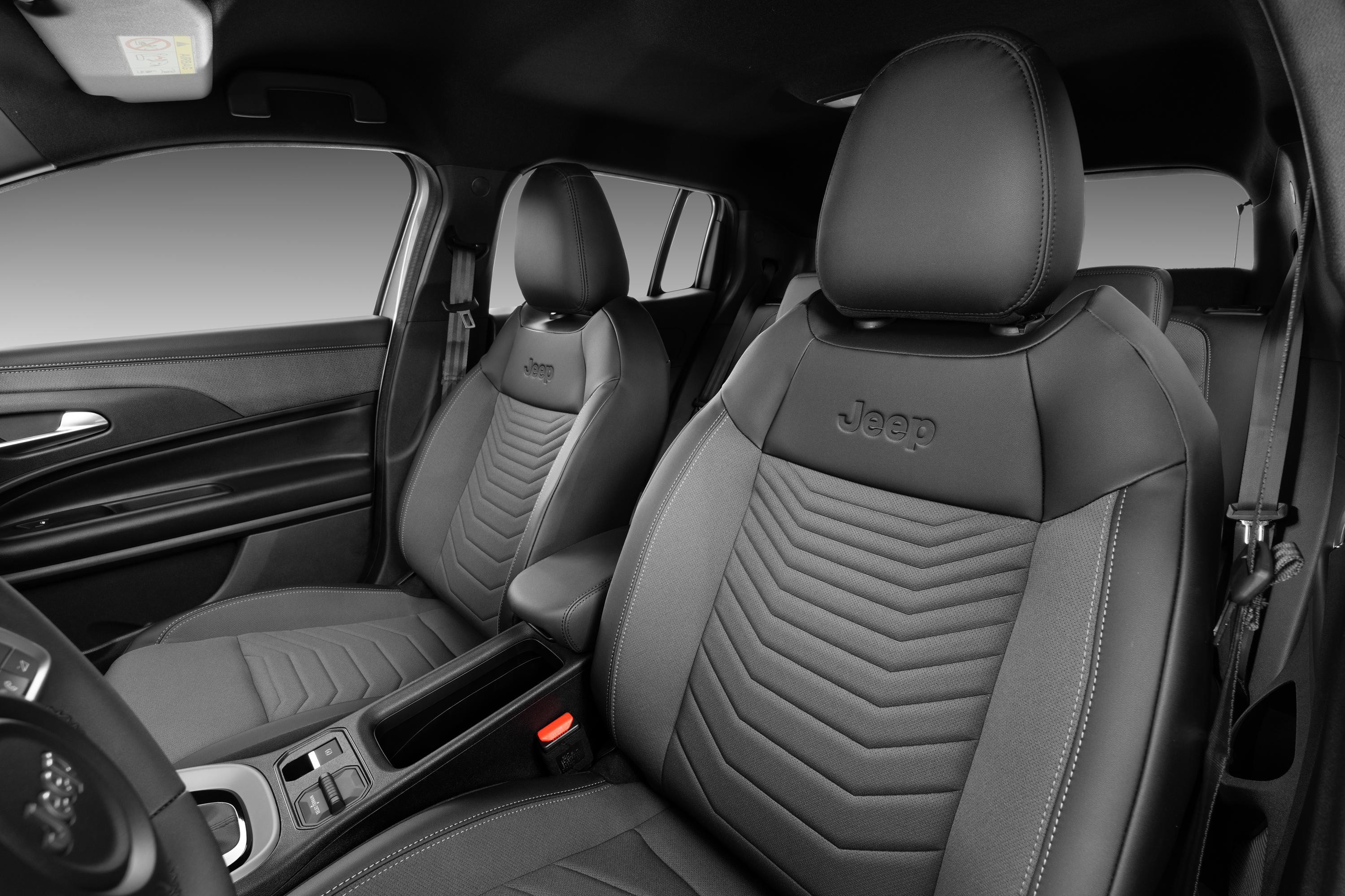 Interior de carro com bancos dianteiros em couro preto e cinza, com costuras e o logotipo Jeep bordado nos encostos de cabe&ccedil;a. O console central e o painel da porta tamb&eacute;m s&atilde;o vis&iacute;veis, com detalhes em preto e cinza, e o volante parcialmente &agrave; esquerda