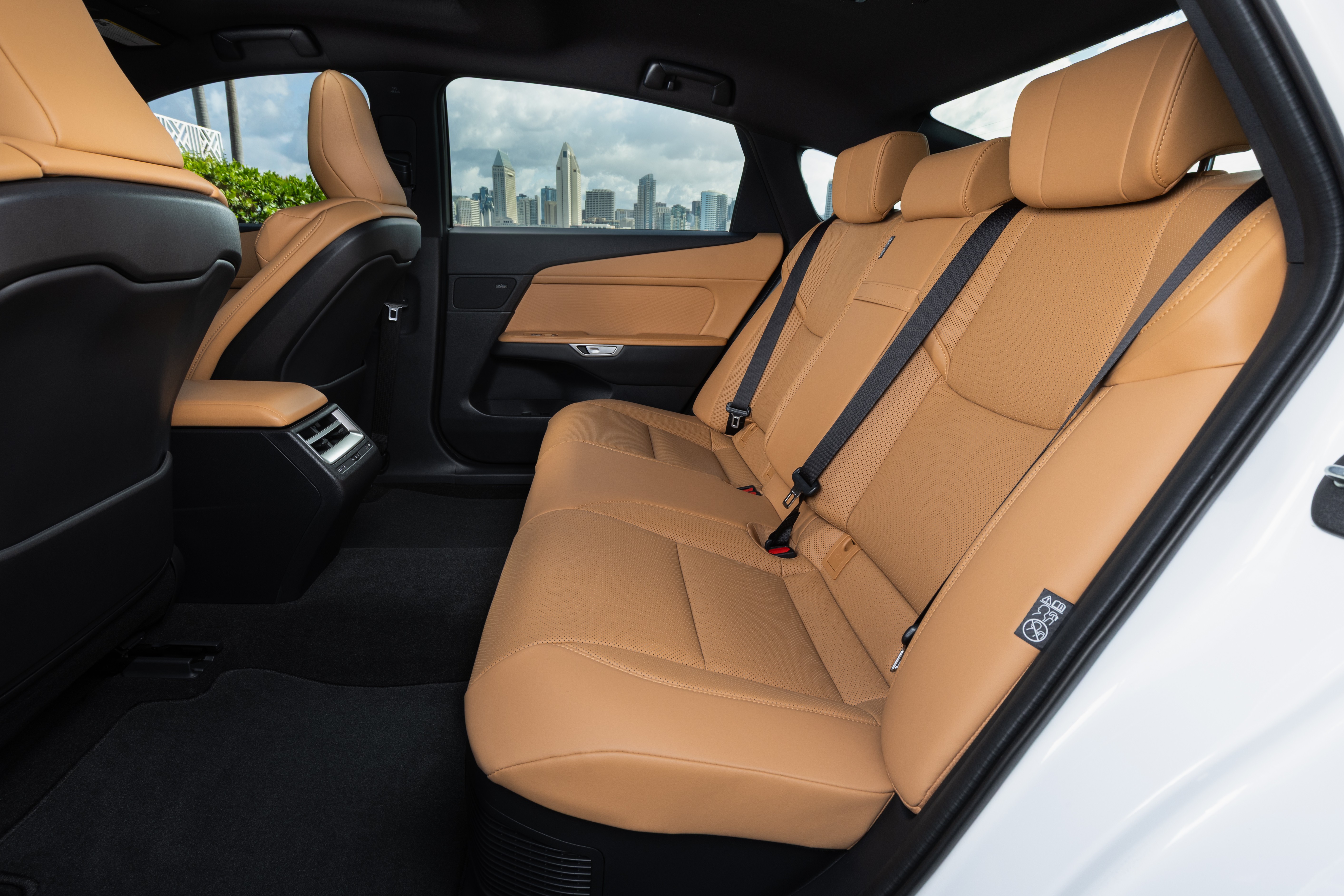 Interior de carro com bancos traseiros de couro marrom-claro e cintos de seguran&ccedil;a pretos. O piso &eacute; forrado com carpete preto. Pela janela, v&ecirc;-se um horizonte urbano com pr&eacute;dios altos sob c&eacute;u nublado