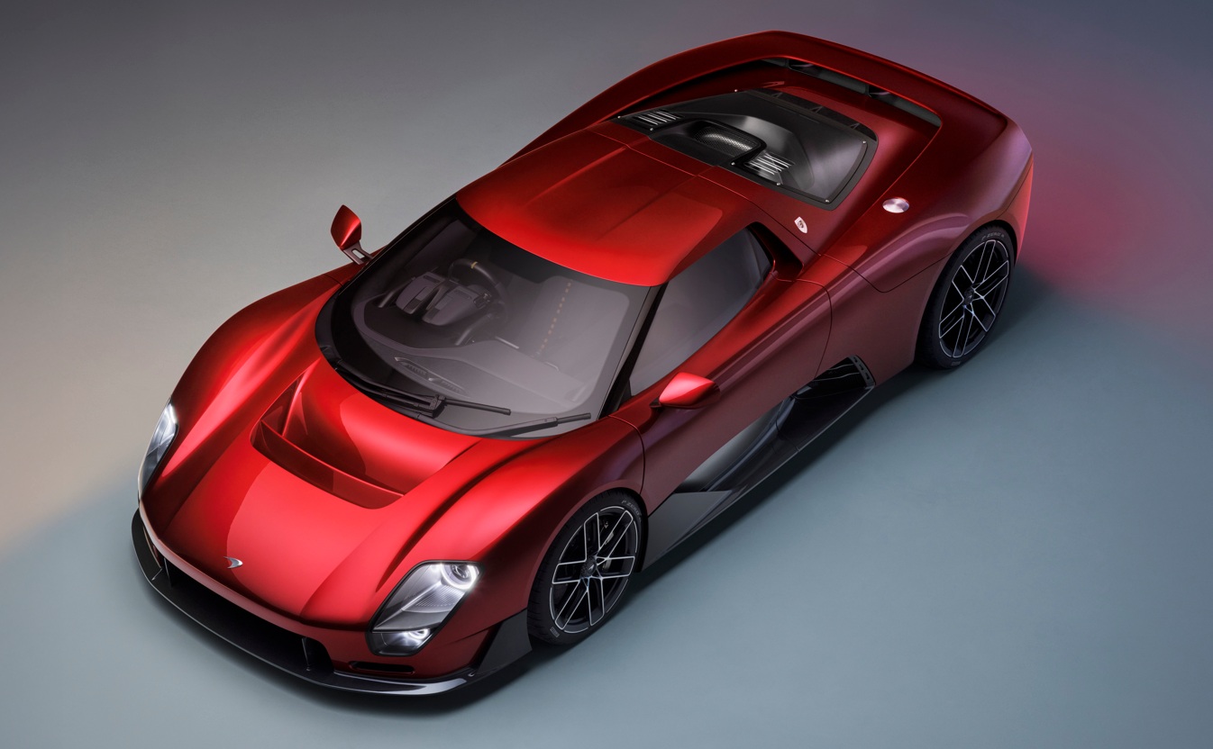 Carro esportivo vermelho met&aacute;lico, visto de cima, com design aerodin&acirc;mico e detalhes em preto fosco, como as rodas e a parte inferior da carroceria. O ve&iacute;culo est&aacute; parado sobre um fundo cinza-azulado, com ilumina&ccedil;&atilde;o suave destacando suas curvas.