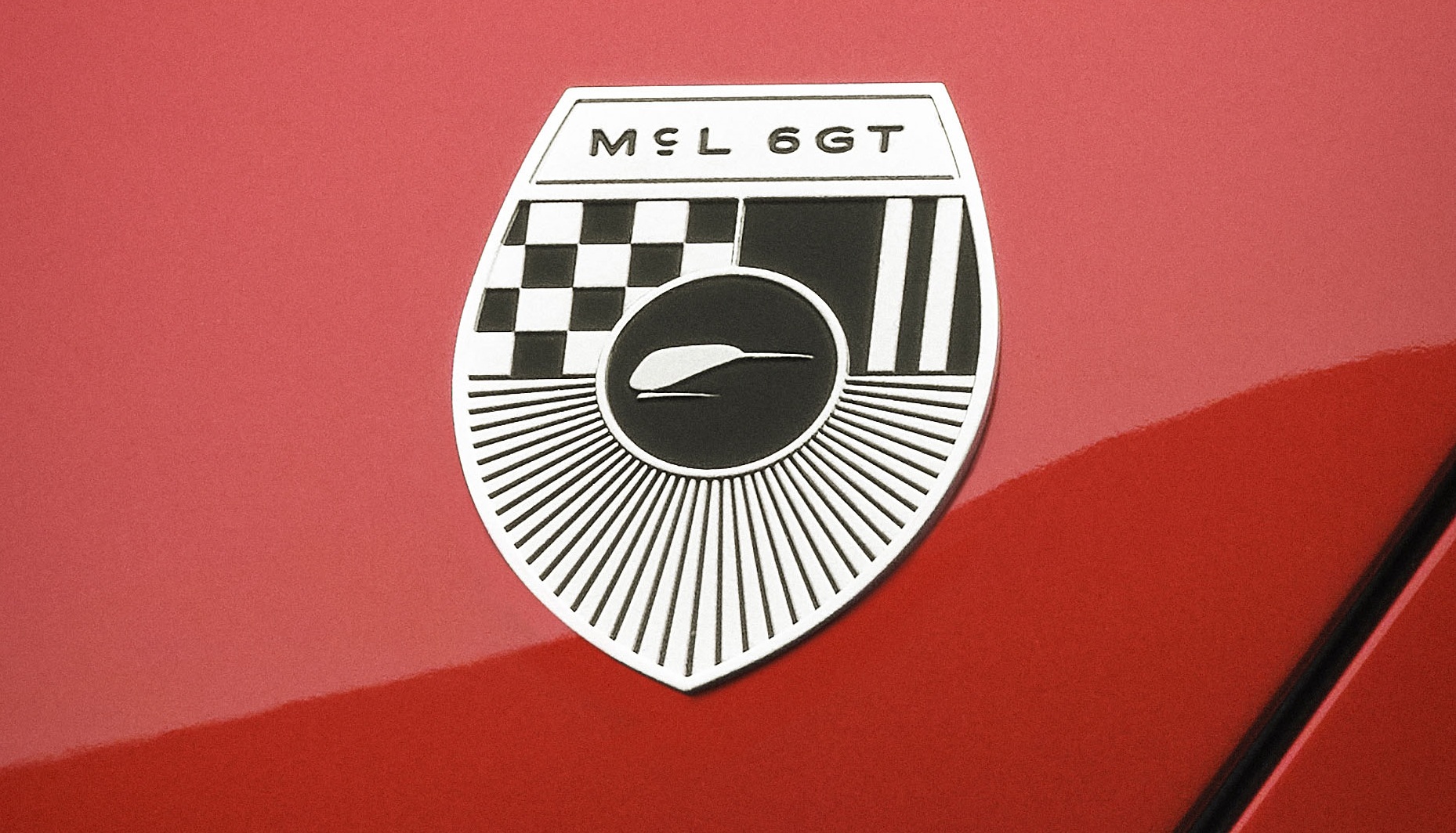 Emblema prateado e preto do carro M&L 6GT em formato de escudo, com um padr&atilde;o xadrez, listras verticais e um desenho de um animal estilizado no centro, sobre uma superf&iacute;cie vermelha brilhante