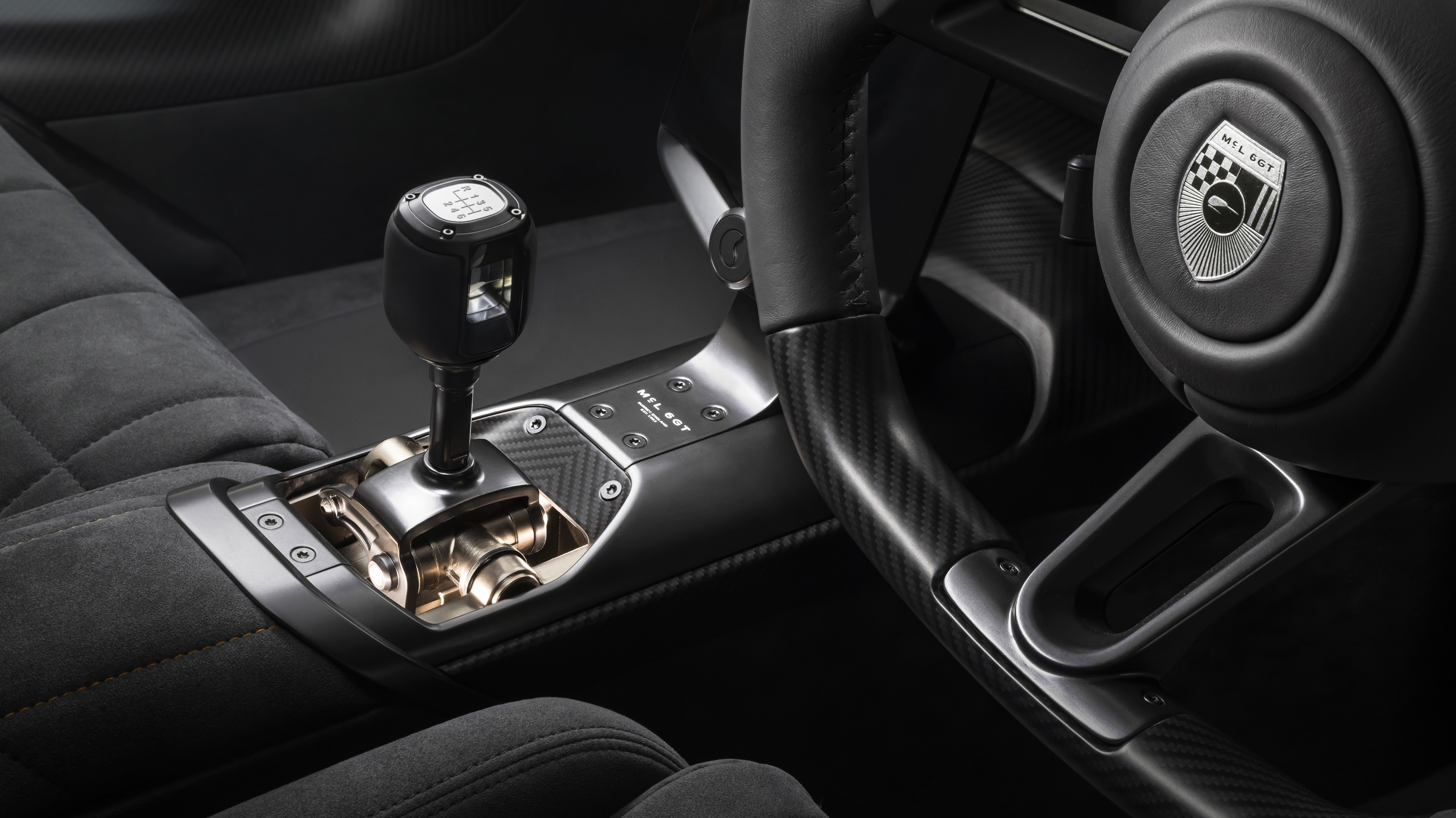 Interior de carro esportivo com c&acirc;mbio manual de seis marchas e r&eacute;, alavanca preta e base dourada exposta. Volante multifuncional com acabamento em fibra de carbono e emblema Mil.ot no centro. Bancos em alc&acirc;ntara cinza com costura laranja. Detalhes em preto e prata, criando um ambiente sofisticado e tecnol&oacute;gico