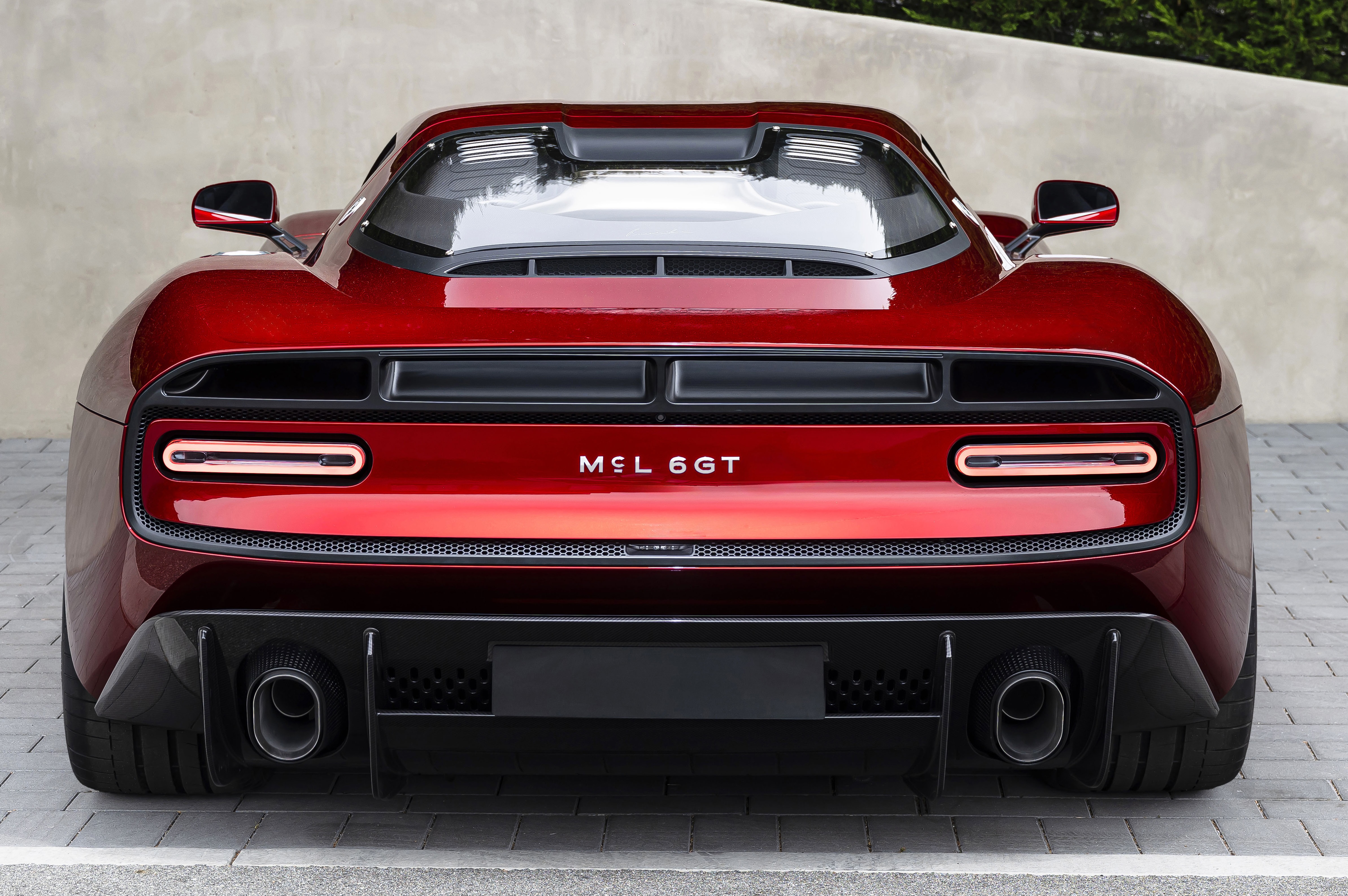 Traseira de um carro esportivo vermelho brilhante, com lanternas horizontais finas e o nome MCL 6GT no centro. Possui duas sa&iacute;das de escapamento circulares e um difusor preto na parte inferior, sobre um piso de blocos cinzas