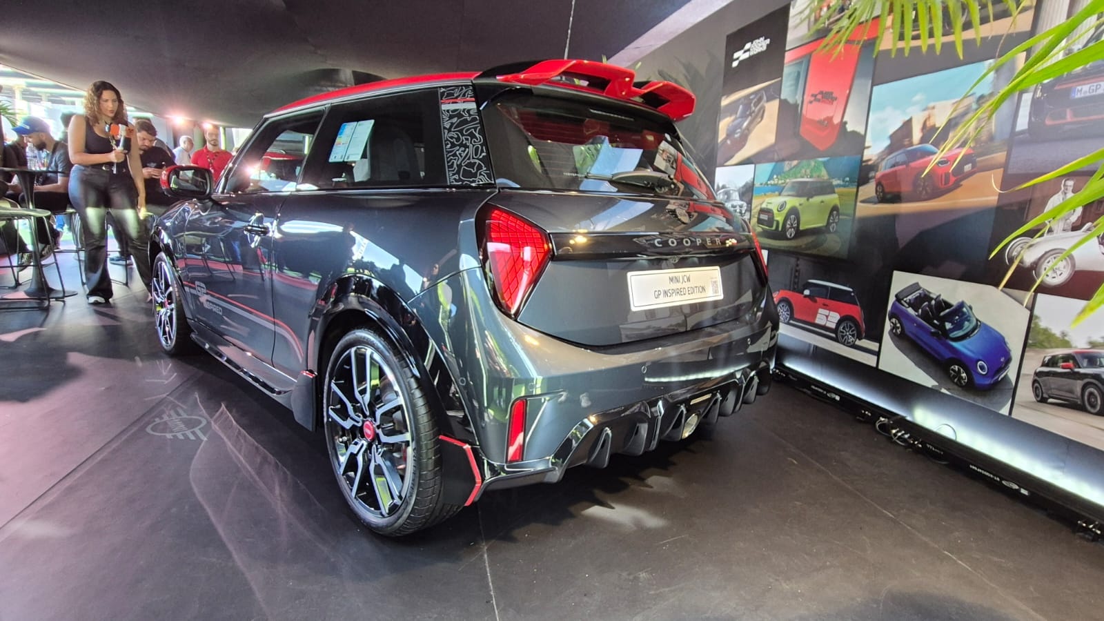 Mini Cooper cinza escuro com teto e aerof&oacute;lio vermelhos, exibido em um evento. Uma mulher de cal&ccedil;a e blusa pretas segura um microfone &agrave; esquerda. Ao fundo, uma parede com fotos de carros coloridos e folhagens verdes no canto direito