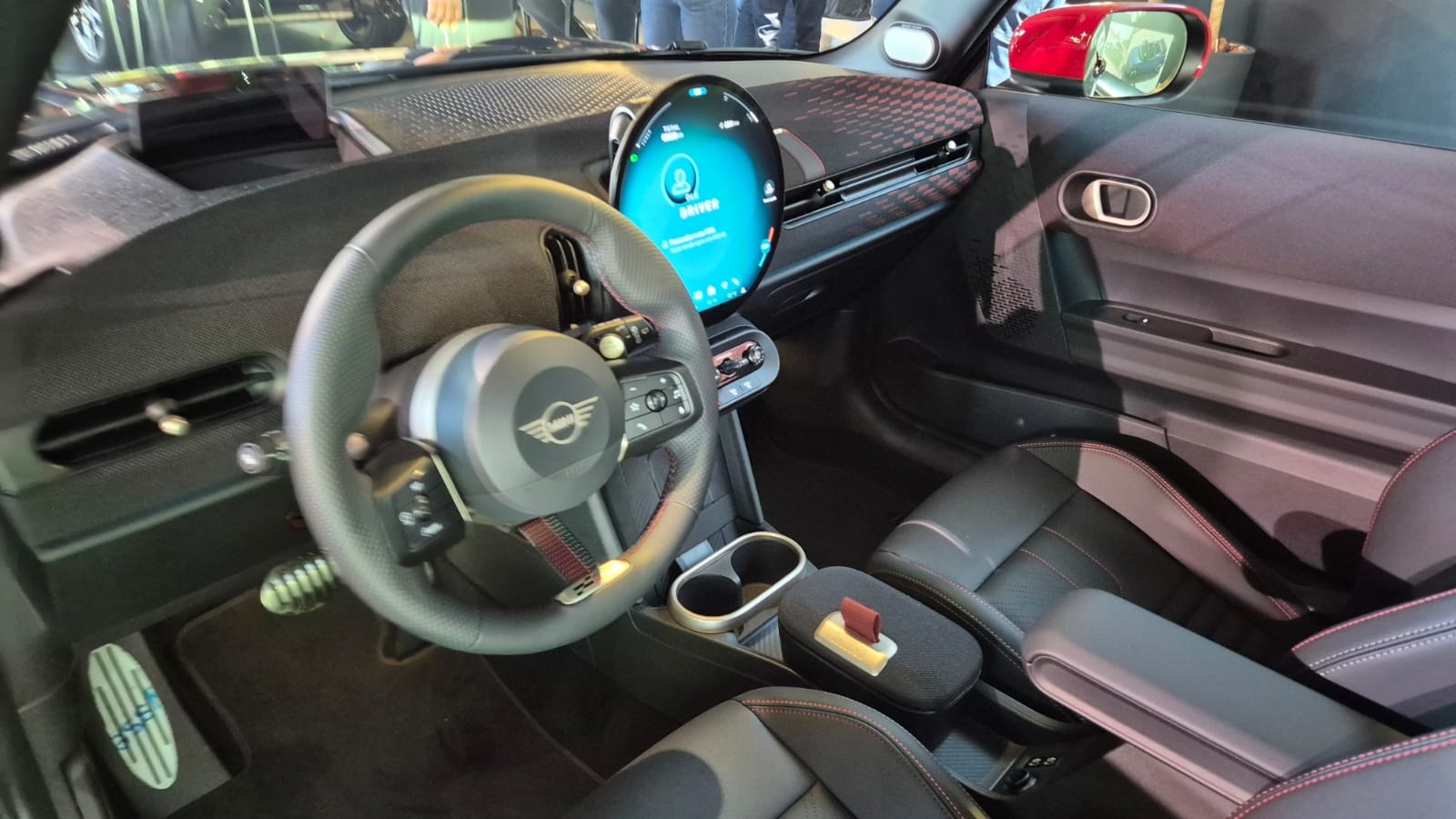 Interior de um carro Mini Cooper com volante preto e o logotipo da marca no centro. Um painel digital redondo e azul exibe &iacute;cones de perfil e configura&ccedil;&otilde;es. O banco do motorista &eacute; preto com detalhes em vermelho, e o console central tem porta-copos e um apoio de bra&ccedil;o. Um retrovisor vermelho &eacute; vis&iacute;vel &agrave; direita