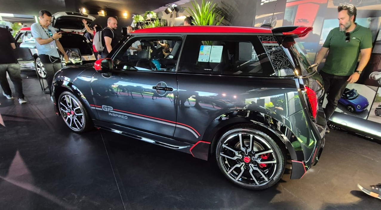 Mini Cooper cinza escuro com teto vermelho e detalhes vermelhos, exposto em um evento. Homens observam o carro e outros ve&iacute;culos ao fundo