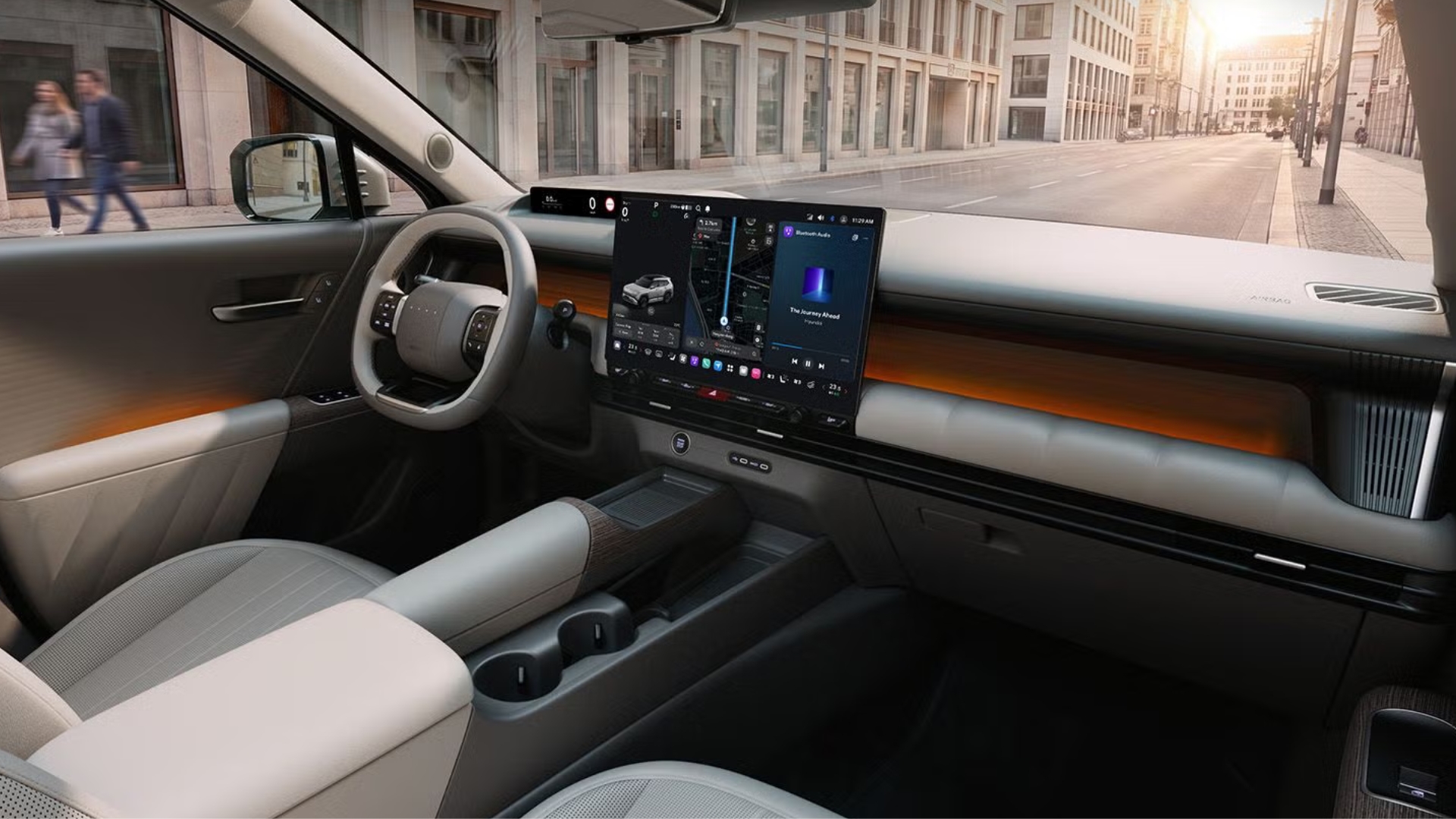 Interior de um carro moderno com volante cinza, tela multim&iacute;dia grande exibindo mapa e m&uacute;sica, e detalhes em madeira clara. O painel tem ilumina&ccedil;&atilde;o ambiente laranja. Ao fundo, uma rua urbana com pr&eacute;dios e duas pessoas caminhando, sob luz solar