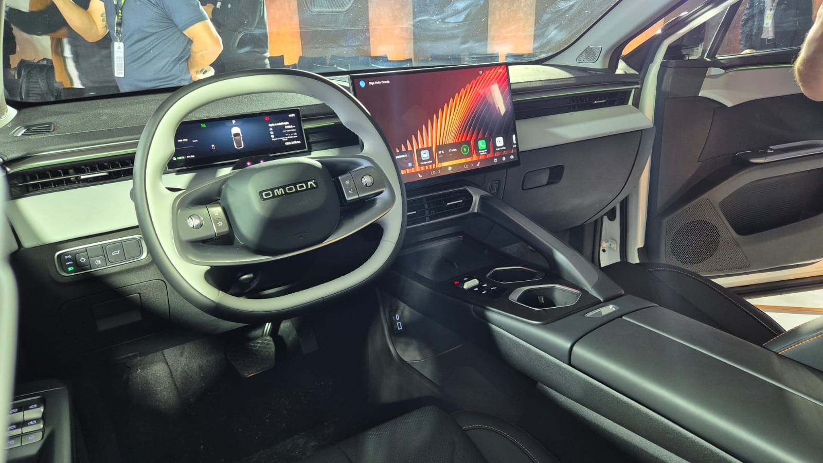 Interior de um carro OMODA com volante multifuncional cinza e branco, painel digital e tela multim&iacute;dia grande exibindo aplicativos. O console central tem porta-copos e acabamento escuro, com detalhes em cinza claro no painel e portas.