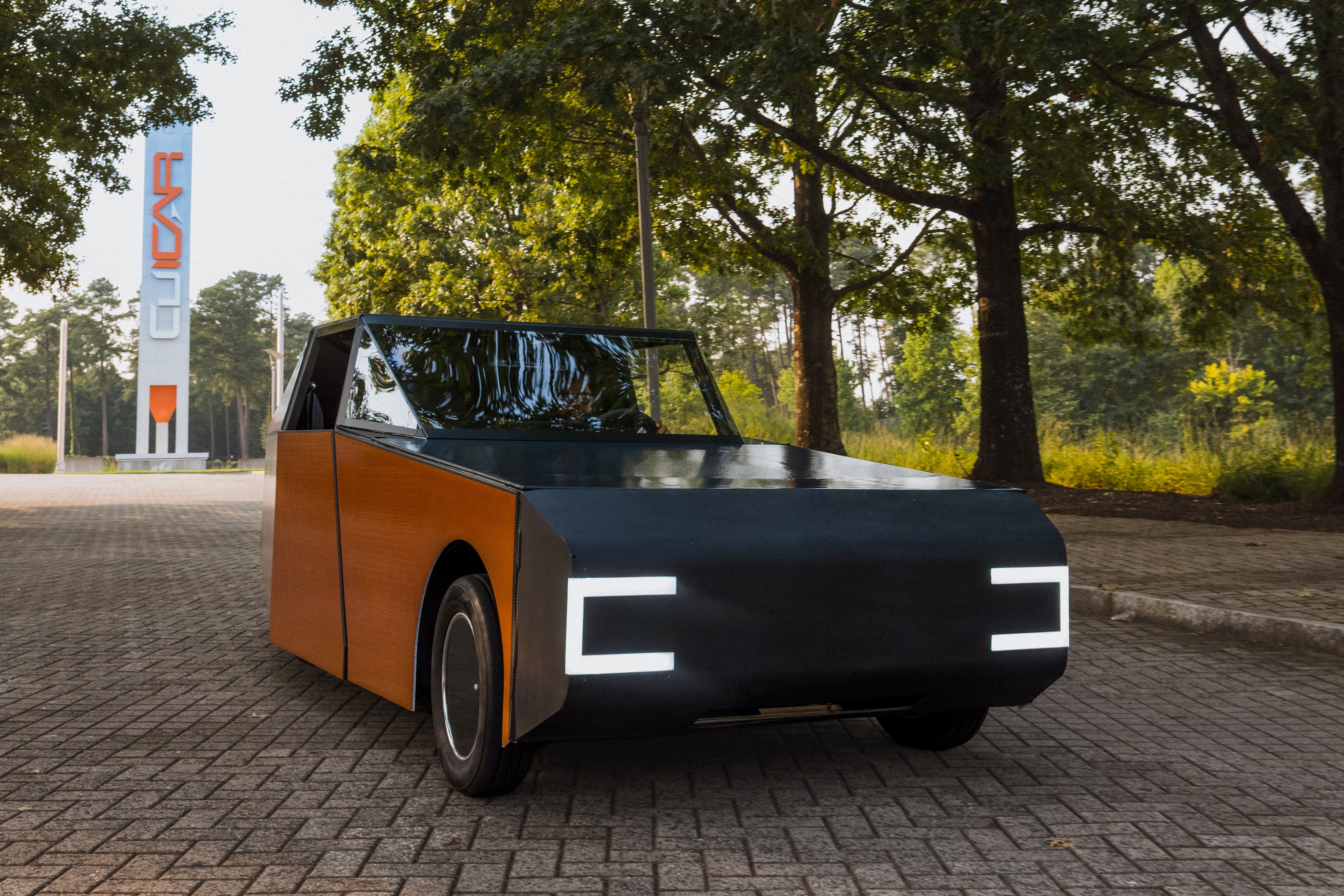 Carro prot&oacute;tipo com design angular, cor preta na frente e marrom nas laterais, com far&oacute;is em formato de C branco. Estacionado em piso de paralelep&iacute;pedos, com &aacute;rvores ao fundo e um monumento branco com a palavra CAR em laranja.