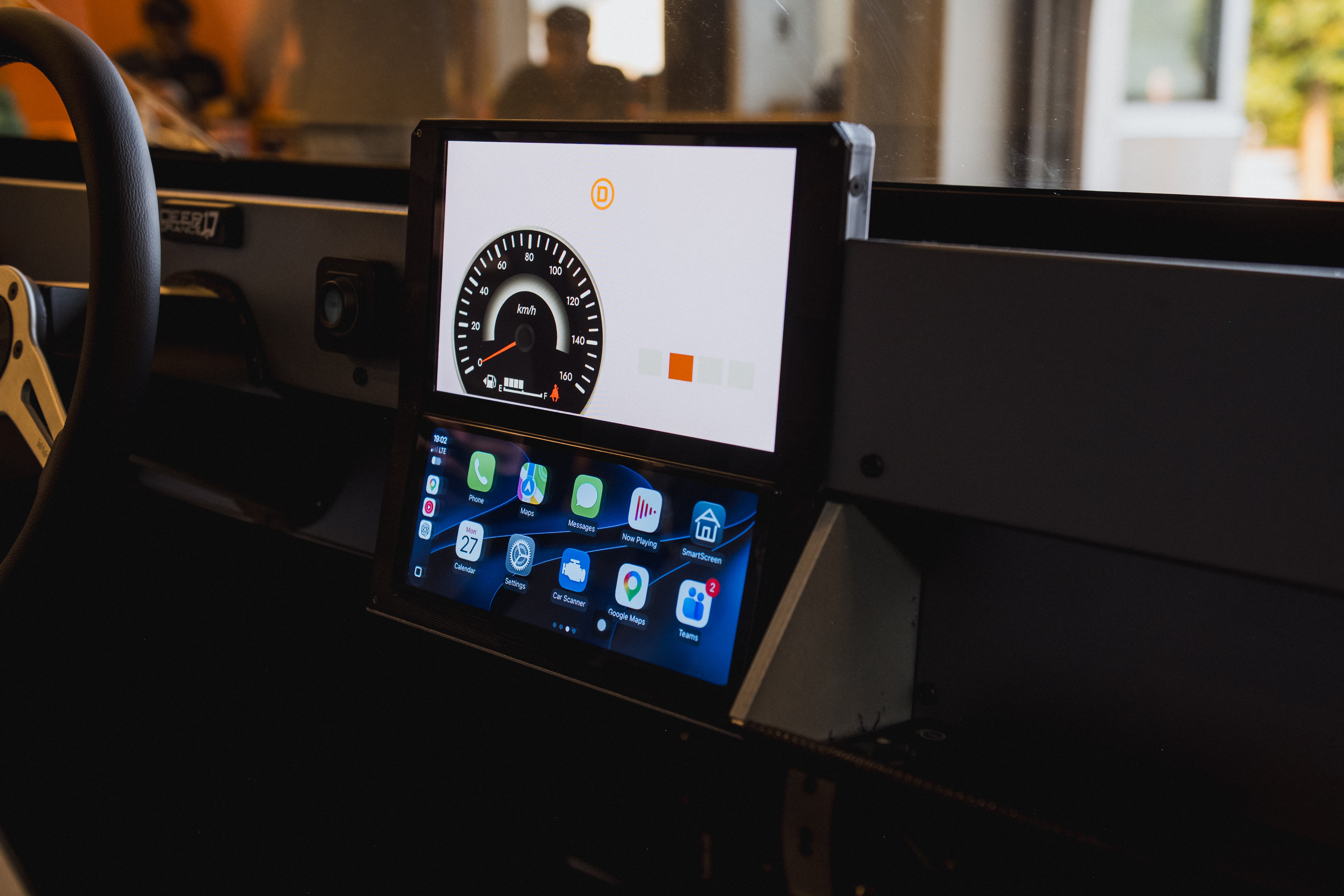 Painel de carro com dois monitores digitais. O superior exibe um veloc&iacute;metro e o inferior, aplicativos de celular em &iacute;cones coloridos. Volante preto &agrave; esquerda.