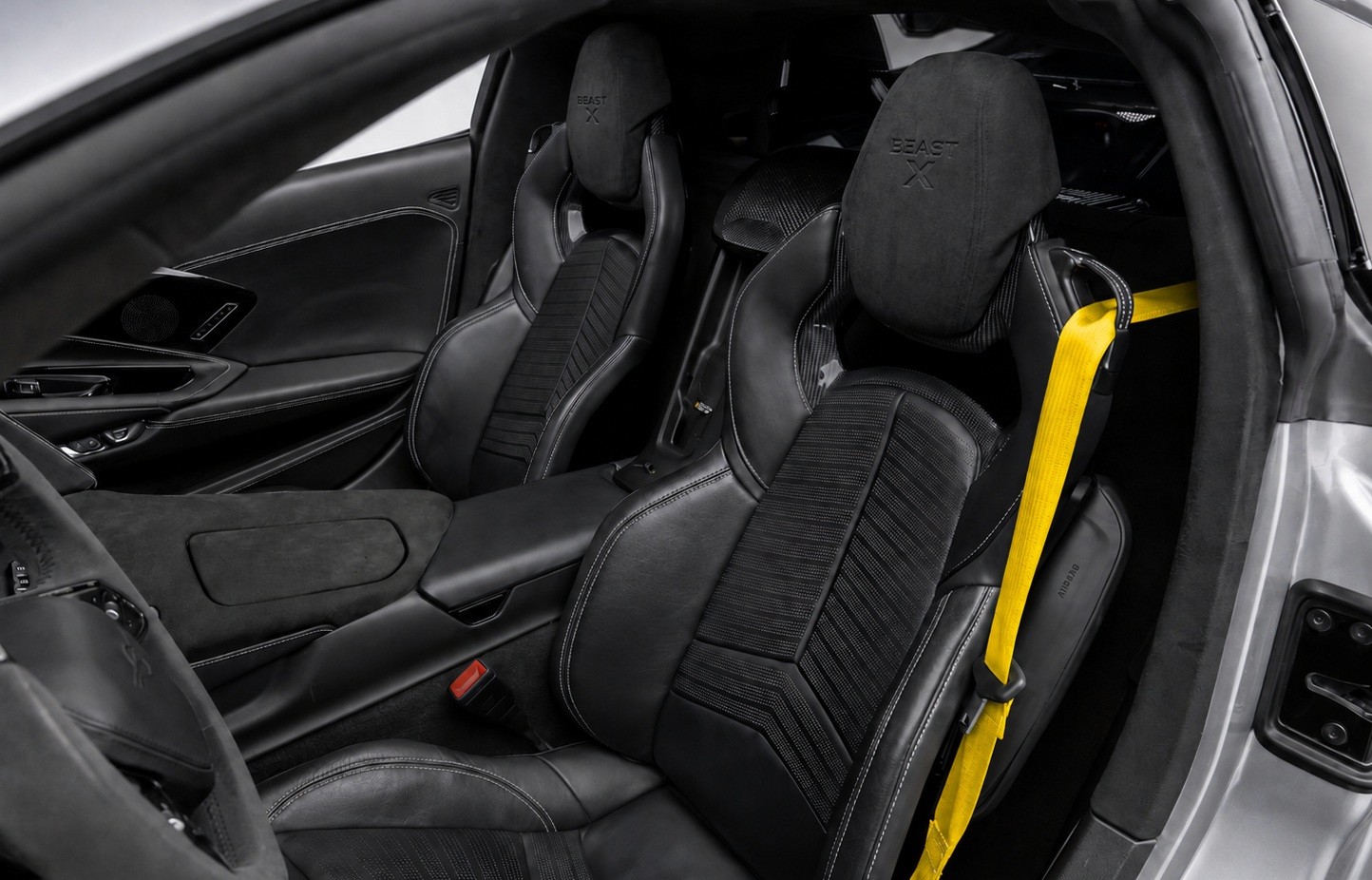 Interior de um carro esportivo com dois bancos dianteiros pretos de couro e tecido, costuras brancas, e o logotipo BEAST X nos encostos de cabe&ccedil;a. O cinto de seguran&ccedil;a do passageiro &eacute; amarelo vibrante, contrastando com o ambiente escuro. O console central e o painel da porta tamb&eacute;m s&atilde;o pretos, com detalhes em costura branca