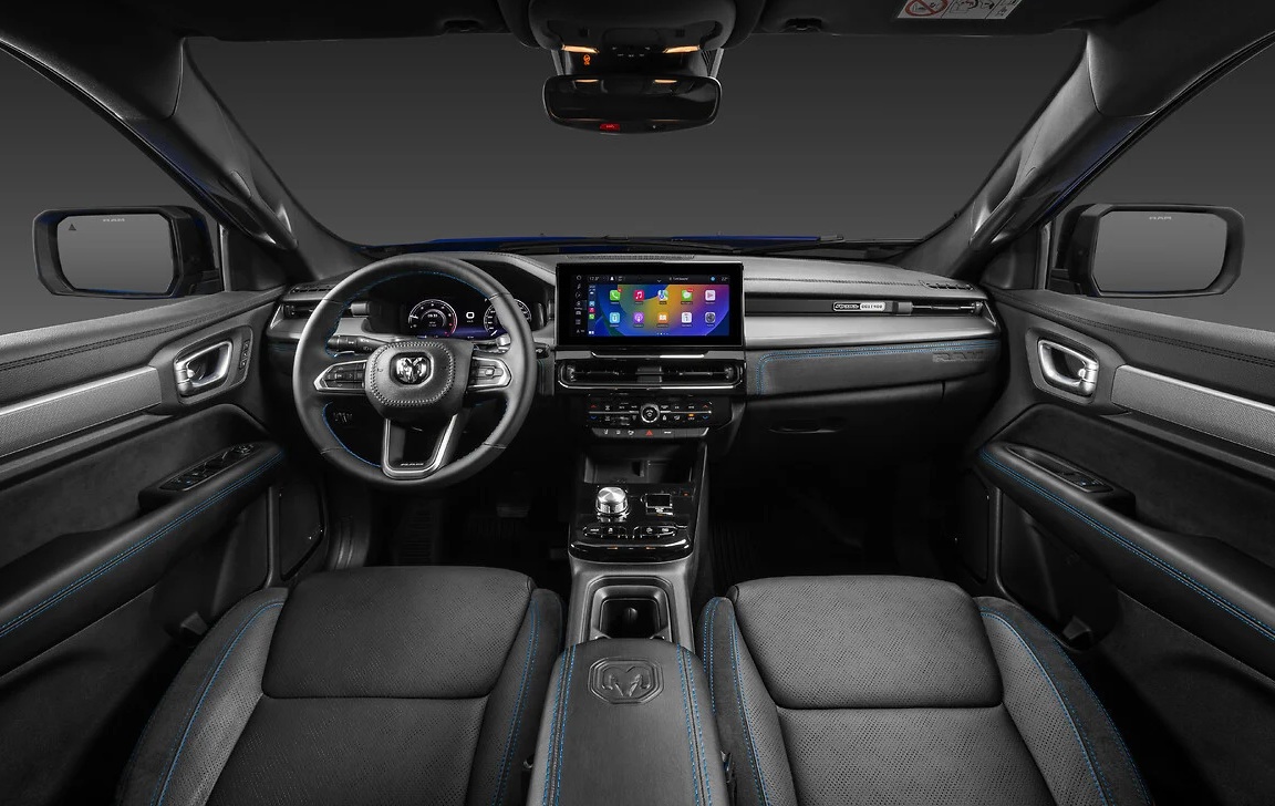 Interior de um carro moderno com bancos de couro escuro e costura azul, volante multifuncional com logo Ram, painel digital e central multim&iacute;dia com tela sens&iacute;vel ao toque exibindo &iacute;cones coloridos