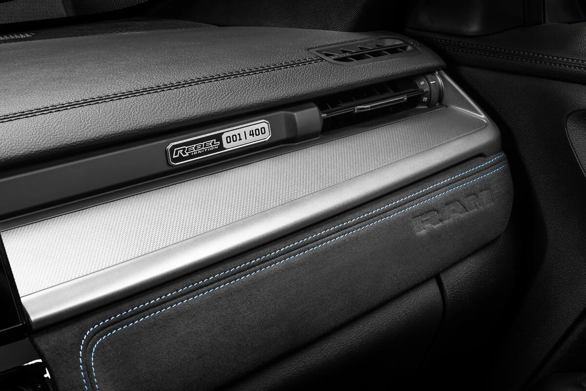 Detalhe do painel de um carro, com acabamento em couro preto e costura azul. Uma placa met&aacute;lica com REBEL IGNITION EDITION 001/400 est&aacute; vis&iacute;vel, acima de uma faixa prateada texturizada. O logotipo RAM est&aacute; gravado no couro abaixo