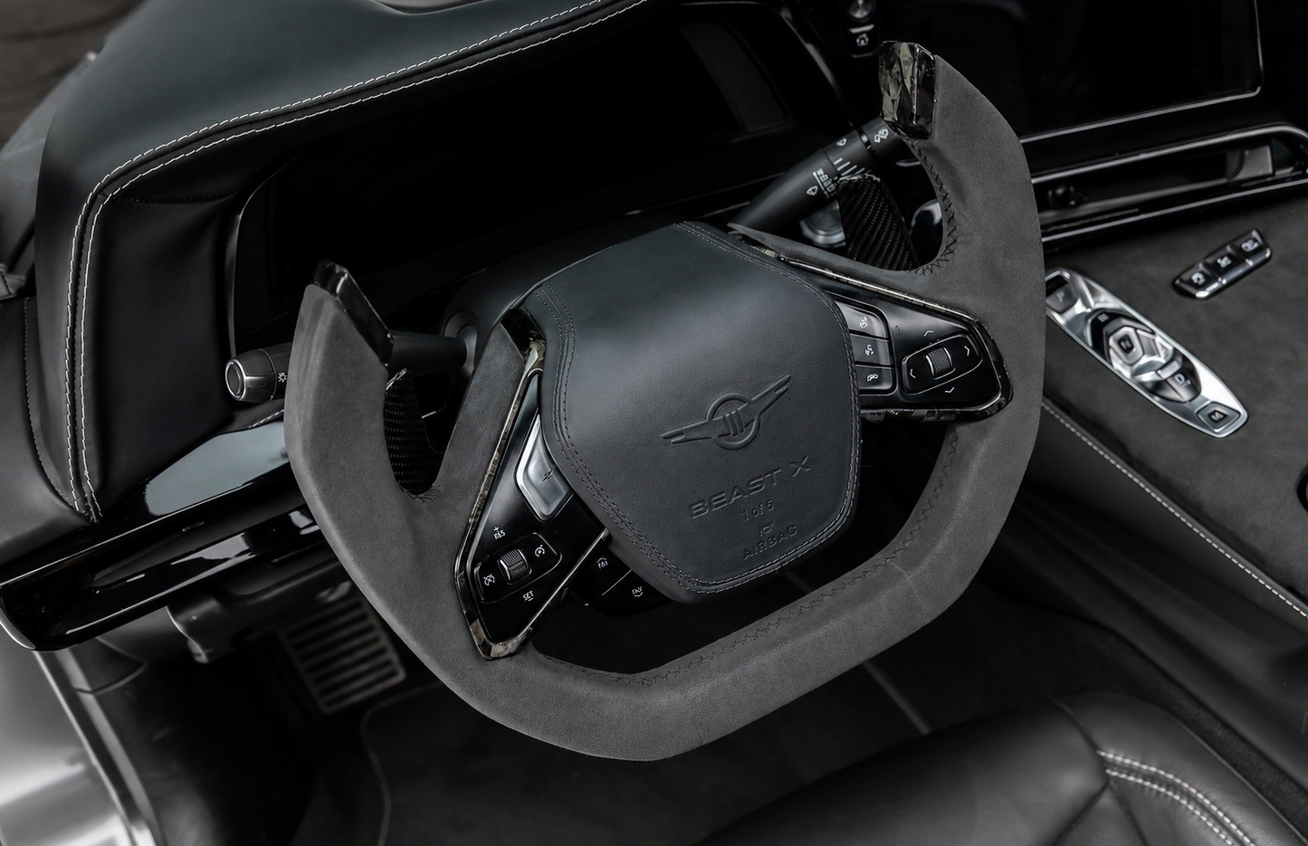 Interior de carro esportivo com volante revestido em camur&ccedil;a escura, costura branca e emblema Beast X no centro. &Agrave; direita, painel com bot&otilde;es de controle e um controle remoto prateado. O ambiente &eacute; luxuoso e moderno, com detalhes em preto e cinza.