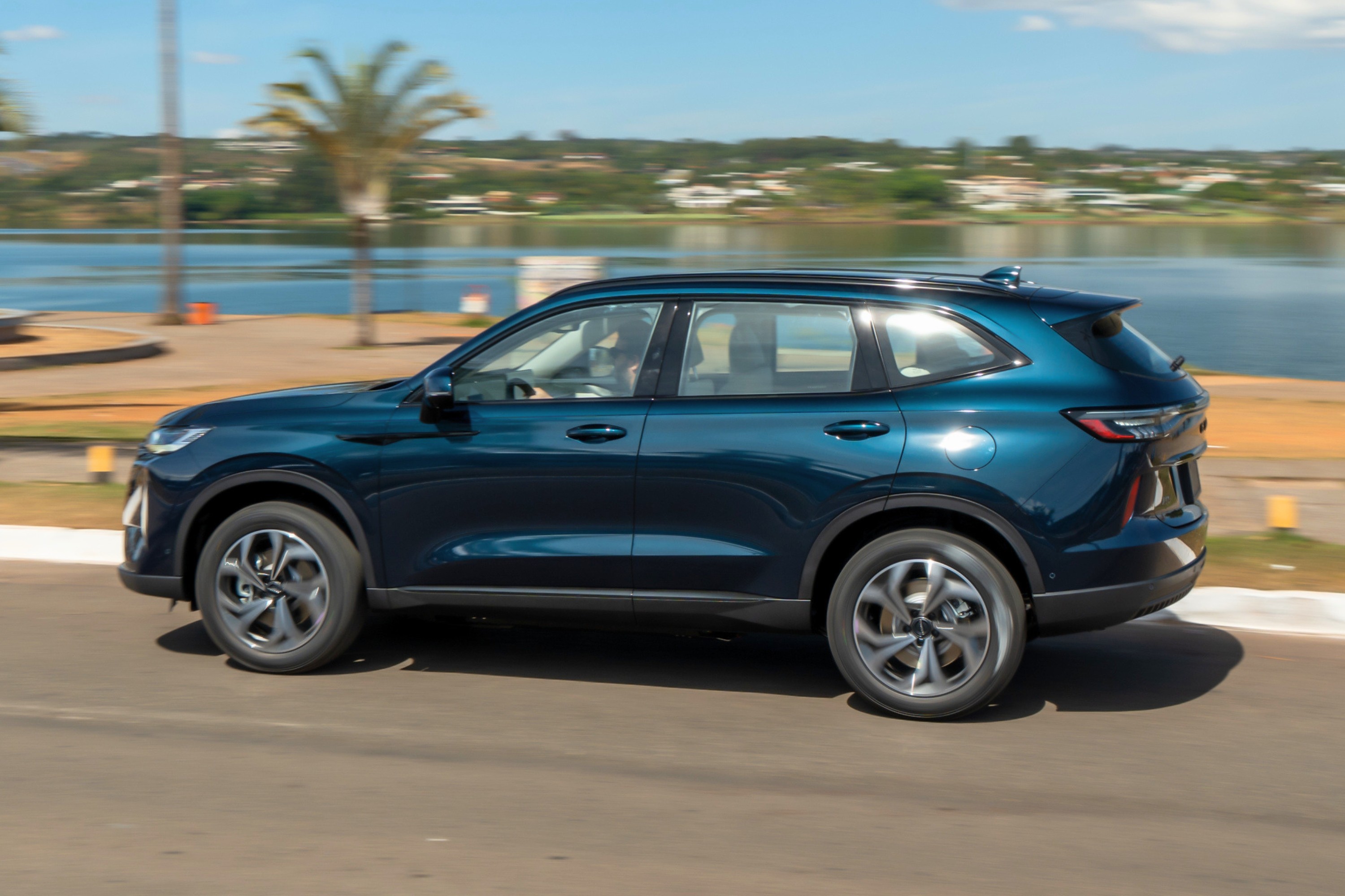 SUV azul-escuro em movimento, visto de perfil, com reflexos de luz solar na lataria. Ao fundo, um lago azul, vegeta&ccedil;&atilde;o e c&eacute;u claro com nuvens esparsas