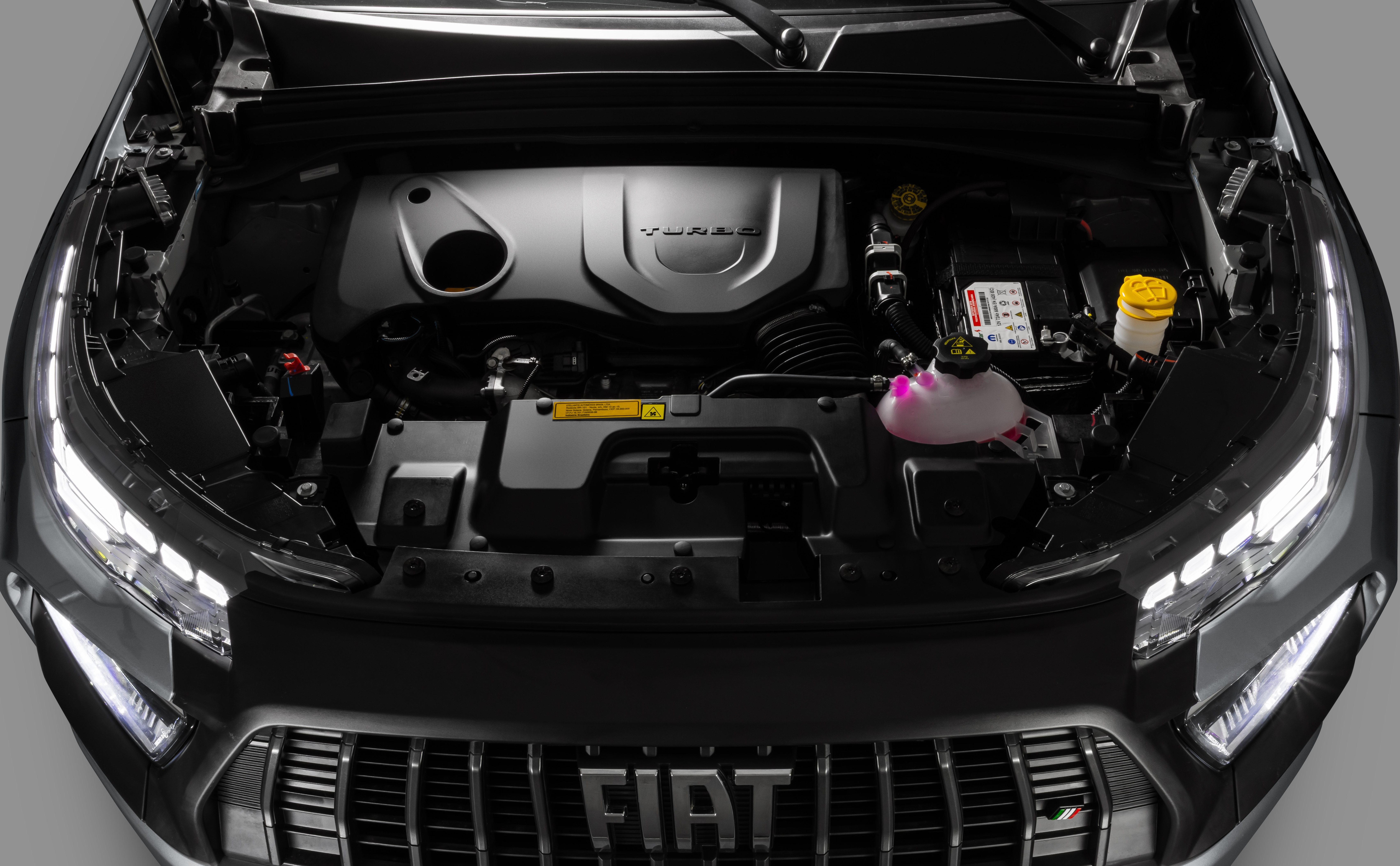 Motor de carro preto com cap&ocirc; aberto, revelando o motor turbo, reservat&oacute;rio de fluido rosa e bateria. A grade frontal com o logo Fiat &eacute; vis&iacute;vel abaixo, com far&oacute;is de LED acesos