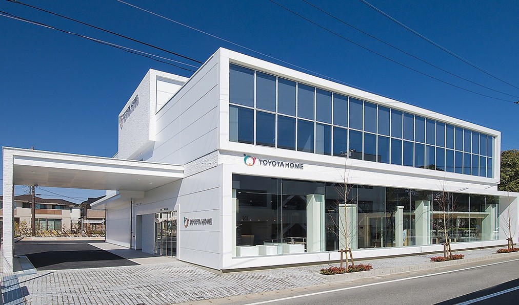 Edif&iacute;cio moderno branco da Toyota Home com grandes janelas espelhadas, uma passarela coberta e estacionamento de paralelep&iacute;pedos, sob c&eacute;u azul claro